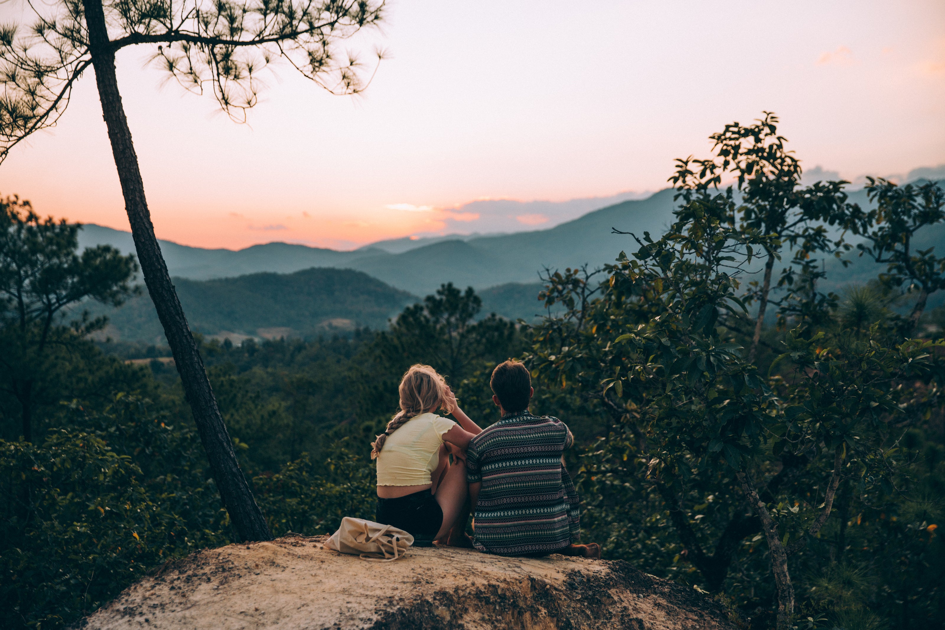 Zwei Personen sitzen auf einem Felsen und blicken in eine weitläufige, bewaldete Berglandschaft bei Sonnenuntergang. Die Szene ist von Bäumen umgeben, und der Himmel zeigt sanfte orange- und rosa Töne.