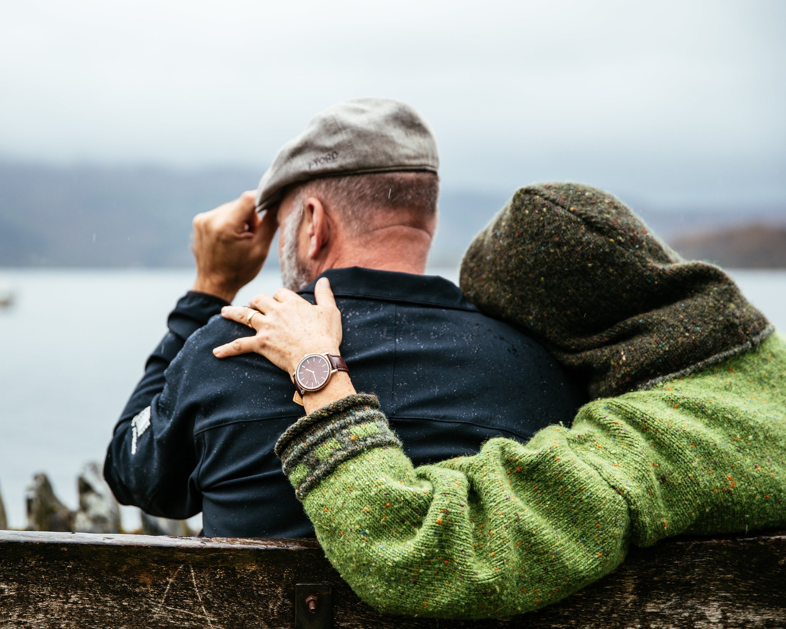 Ein Paar sitzt auf einer Holzbank an einem See. Eine Person trägt einen grünen Pullover und legt den Arm um die andere Person, die eine graue Mütze trägt. Im Hintergrund ist eine ruhige, neblige Landschaft mit Wasser und einem Boot zu sehen.