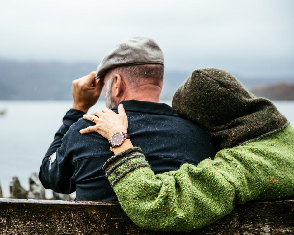 Ein Paar sitzt auf einer Holzbank an einem See. Eine Person trägt einen grünen Pullover und legt den Arm um die andere Person, die eine graue Mütze trägt. Im Hintergrund ist eine ruhige, neblige Landschaft mit Wasser und einem Boot zu sehen.