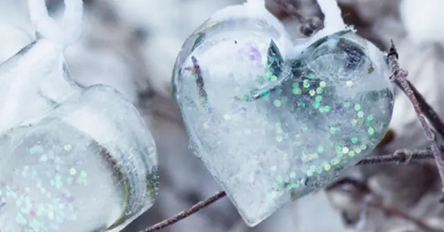 Transparente, herzförmige Eiskristalle mit eingeschlossenem Glitzer, hängend an Ästen in einer winterlichen Umgebung.