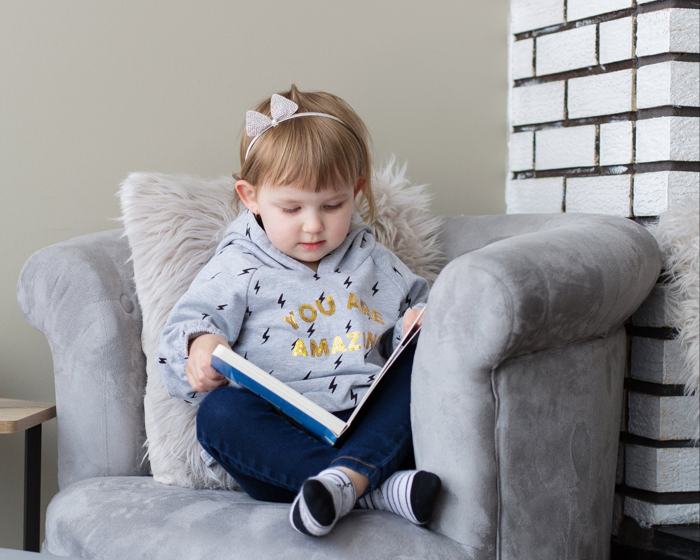 Ein kleines Kind sitzt in einem grauen Sessel, umgeben von weichen Kissen, und liest ein aufgeschlagenes Buch. Im Hintergrund ist eine helle Wand und ein kleiner Beistelltisch mit einer schwarzen Tasse zu sehen. Rechts im Bild befindet sich eine weiß gemauerte Wand.