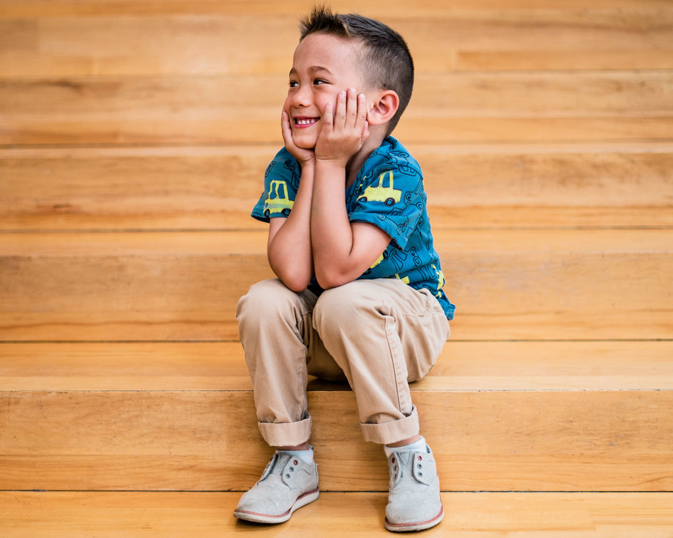 A child sits on wooden stairs, resting their hands under their chin. They are wearing a blue shirt with a car pattern, beige pants, and gray shoes with rolled cuffs.