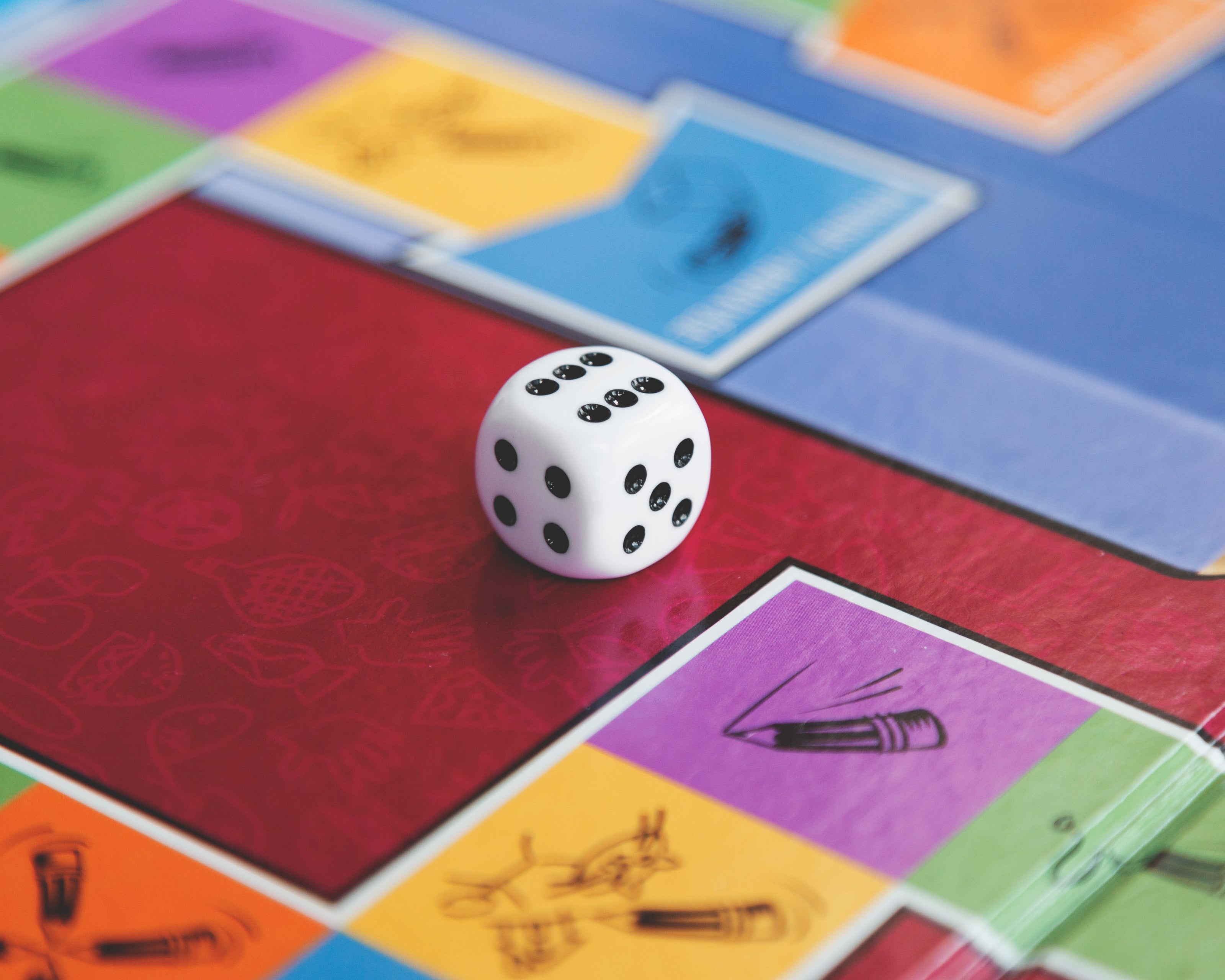 Ein Spielbrett mit bunten Feldern in Rot, Gelb, Blau, Lila und Grün. Auf dem roten Spielfeld liegt ein weißer Würfel mit schwarzen Punkten, der die Zahl sechs zeigt. Die Felder enthalten kleine Illustrationen, wie einen Stift oder eine Figur, die eine Bewegung darstellt. Die Szene deutet auf ein Brettspiel hin.