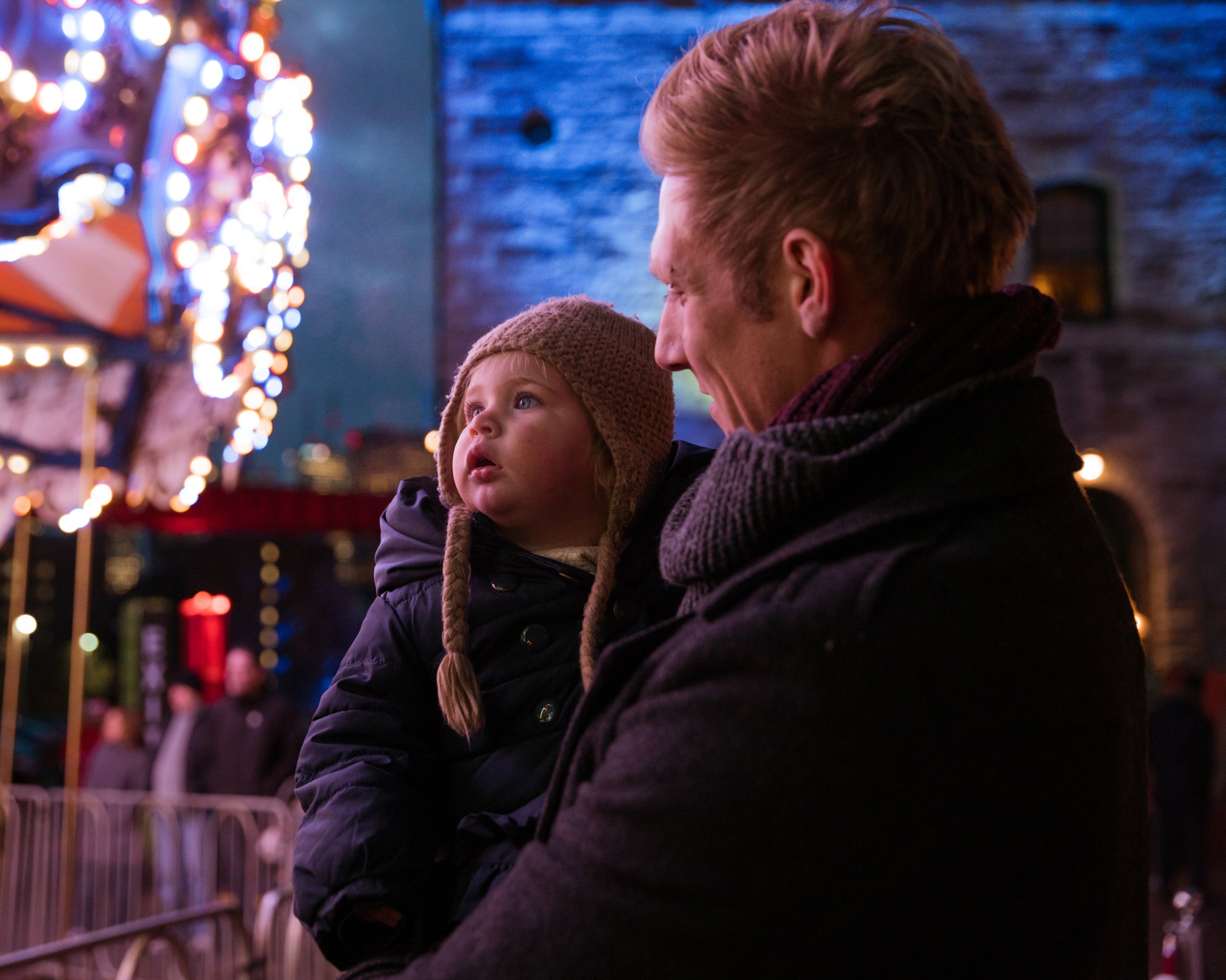 A person holds a child dressed in a warm coat and knit hat during an evening outing. In the background, festive lights and a carousel are visible, along with a building facade and blurred figures.