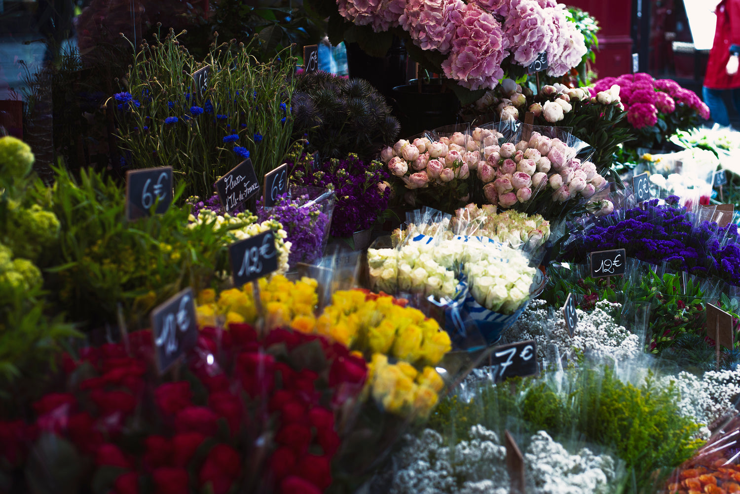 Ein farbenfroher Blumenstand mit einer großen Auswahl an frischen Blumen, darunter rote Rosen, gelbe und weiße Blumen, Lavendel sowie rosa Hortensien. Die Blumen sind in Bündeln arrangiert, mit kleinen Preisschildern versehen, die Beträge wie 6 € und 7 € anzeigen.