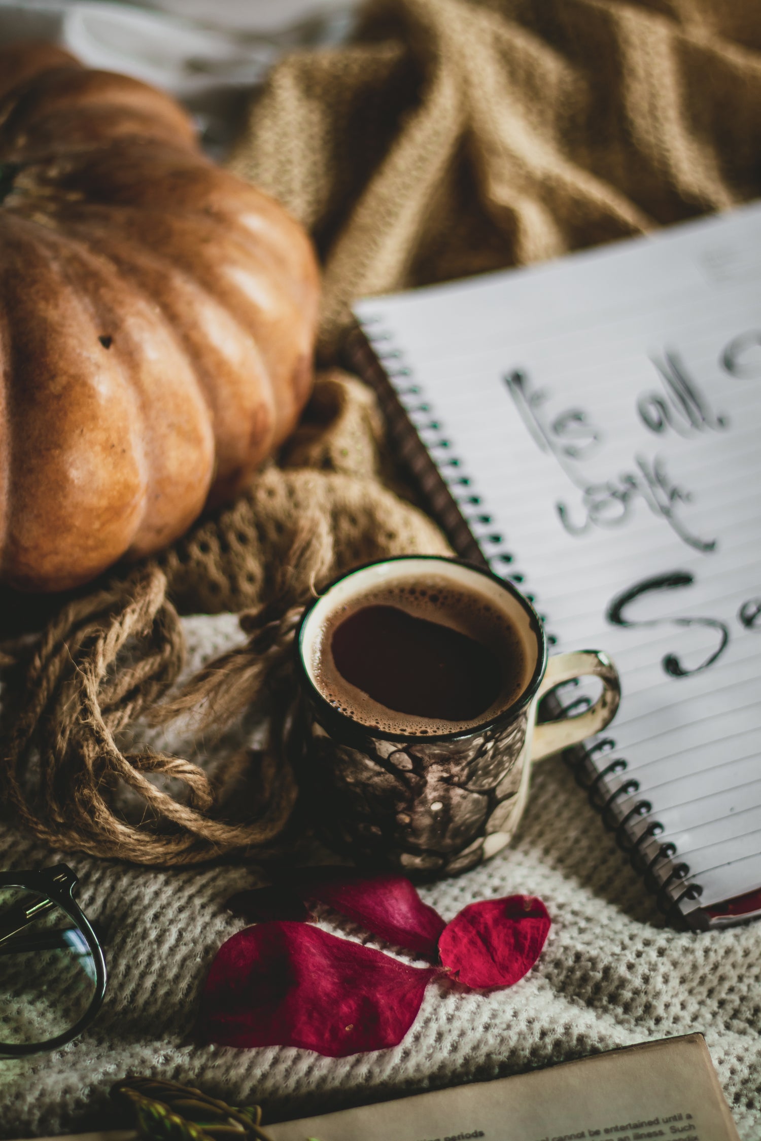 Ein stimmungsvolles Herbst-Arrangement: Im Vordergrund steht eine Tasse Kaffee in einer rustikalen Tasse mit Holzoptik, umgeben von einem roten Herbstblatt, einem Stück grober Schnur und einer Lupe. Im Hintergrund liegen ein großer Kürbis, ein beiges Strickgewebe und ein Spiralblock mit handgeschriebener Notiz, die teilweise lesbar ist ('Is all... safe?').