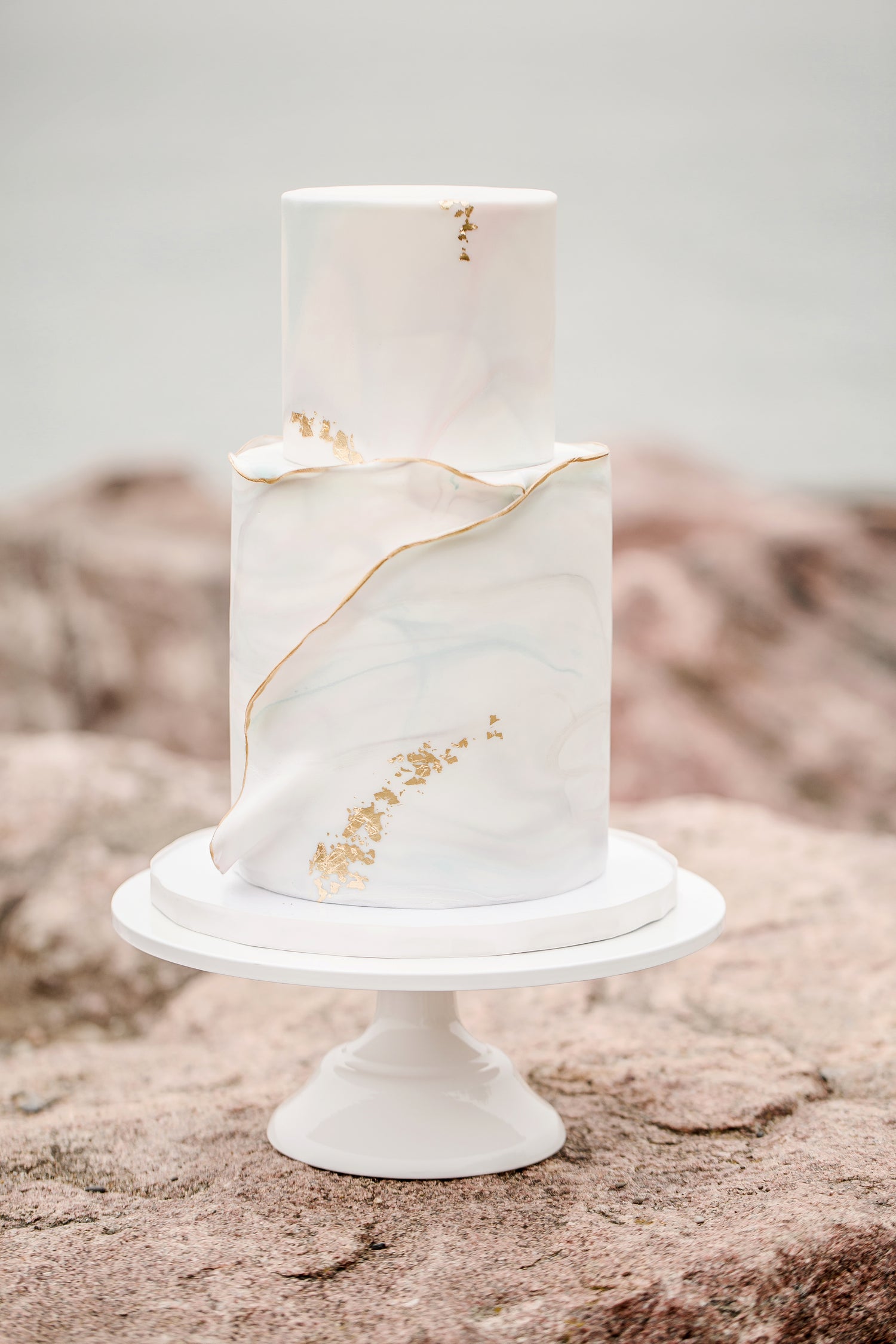 Zweistöckige Hochzeitstorte mit marmoriertem, weißem Fondant, verziert mit goldenen Akzenten und feinen Linien. Die Torte steht auf einer weißen Tortenplatte vor einem natürlichen Hintergrund mit sandfarbenen Felsen und unscharfem Himmel.