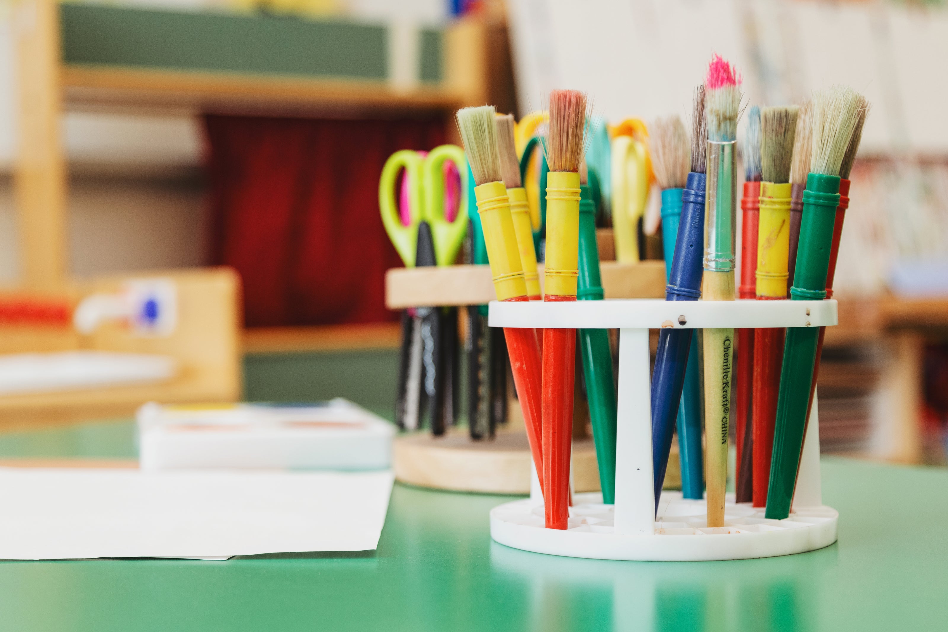 "Ein weißer Stifthalter mit bunten Pinseln und Stiften auf grünem Tisch, unscharfer Hintergrund mit Scheren und rotem Vorhang.