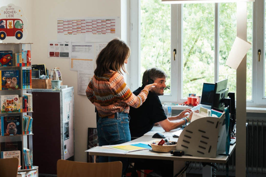Ein heller Arbeitsraum mit großen Fenstern und Blick ins Grüne. Links stehen zwei Bücherständer mit Kinderbüchern, an der Wand Notizen. Zwei Personen arbeiten an einem Schreibtisch mit Büchern und Büromaterial.