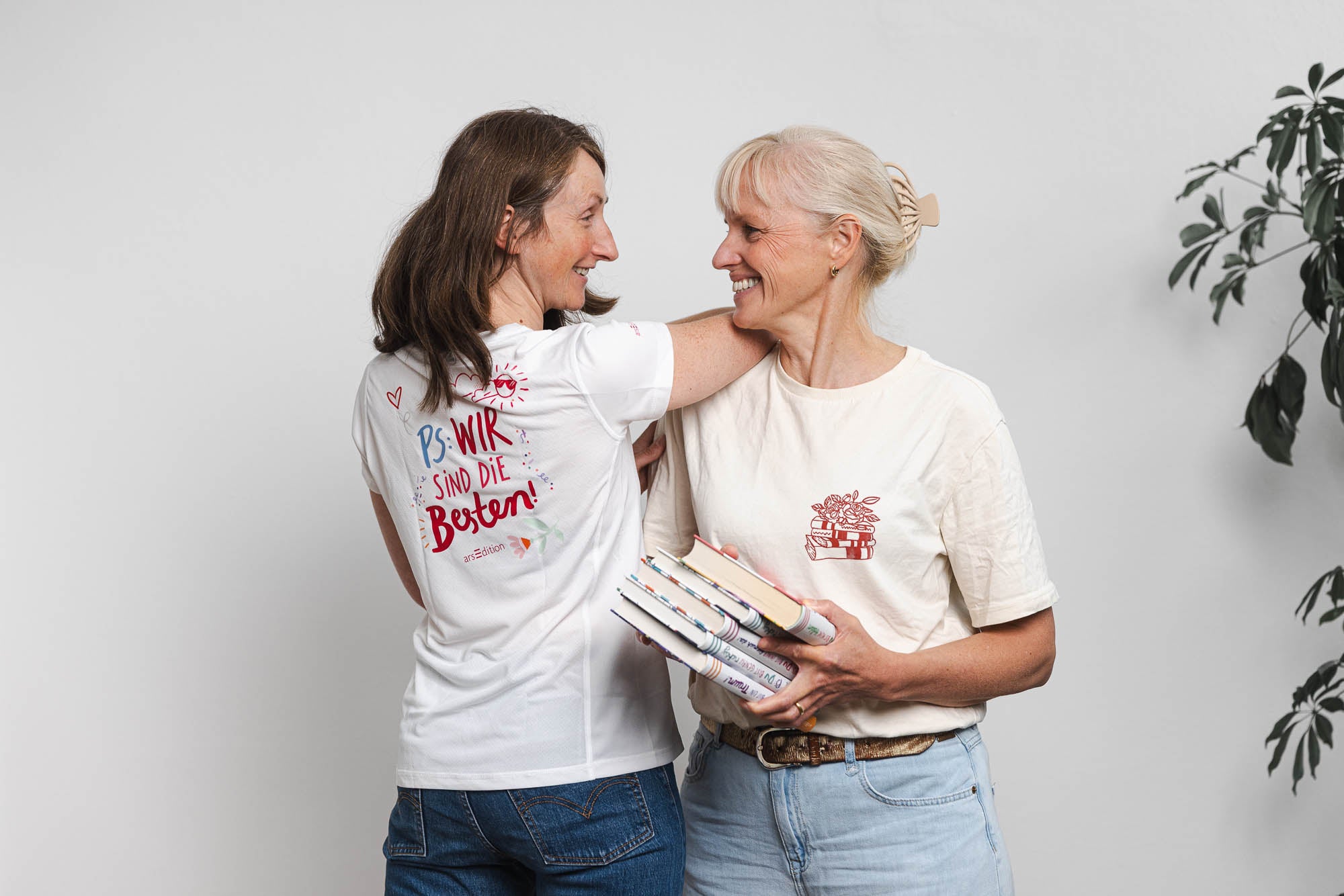 Zwei Personen stehen nebeneinander vor einem neutralen, weißen Hintergrund. Die linke Person trägt ein weißes T-Shirt mit der Rückenaufschrift „PS: WIR SIND DIE BESTEN!“ in bunten Buchstaben und kombiniert es mit einer blauen Jeanshose. Die rechte Person trägt ein cremefarbenes T-Shirt mit einer roten Illustration von gestapelten Büchern und hält mehrere Bücher in den Händen. Rechts am Bildrand ist ein Teil einer grünen Zimmerpflanze sichtbar.