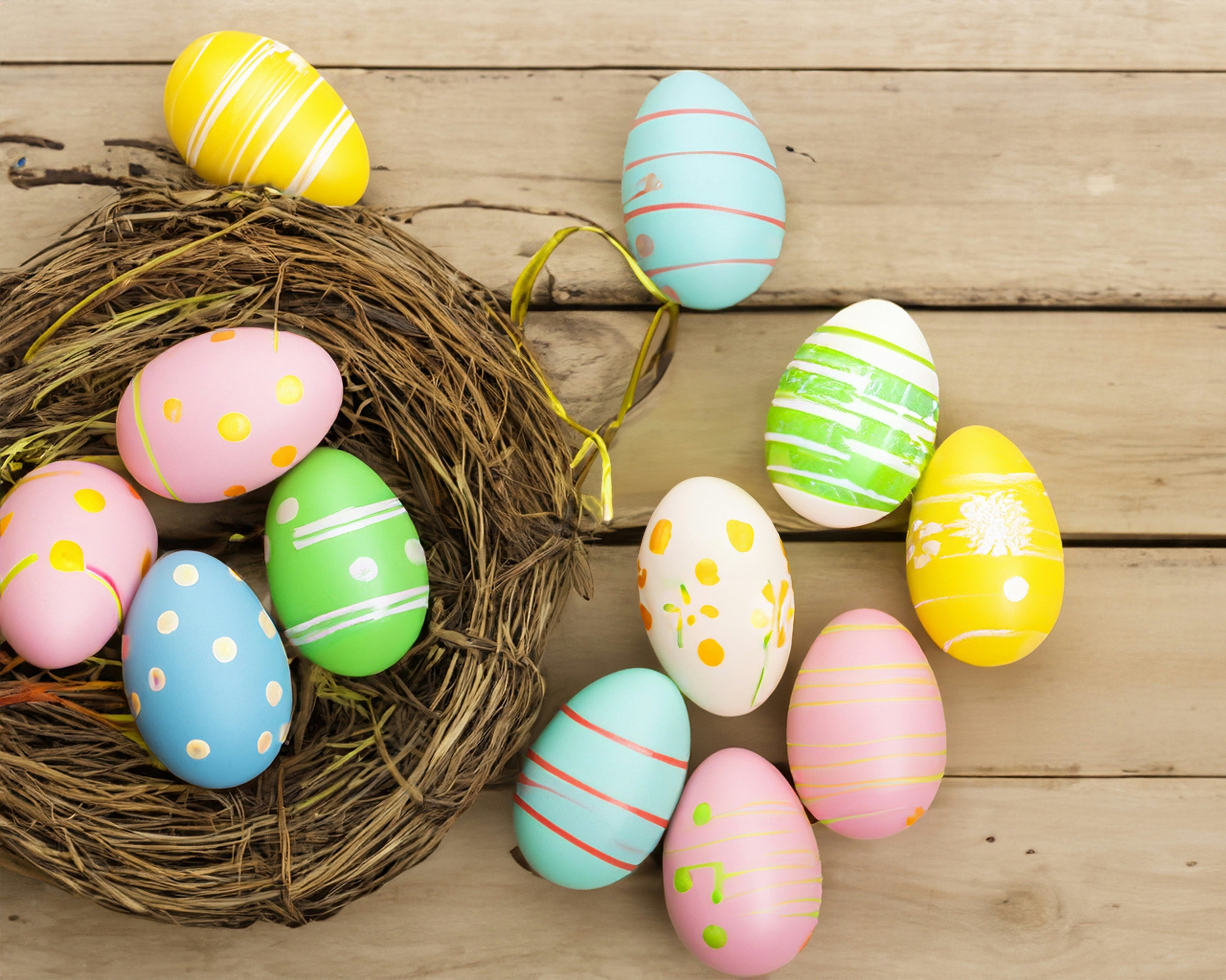 Ein Arrangement aus bunt bemalten Ostereiern in Pastellfarben mit verschiedenen Mustern wie Punkten und Streifen. Die Eier liegen teils in einem dekorativen Nest und teils auf einer Holzoberfläche verteilt.