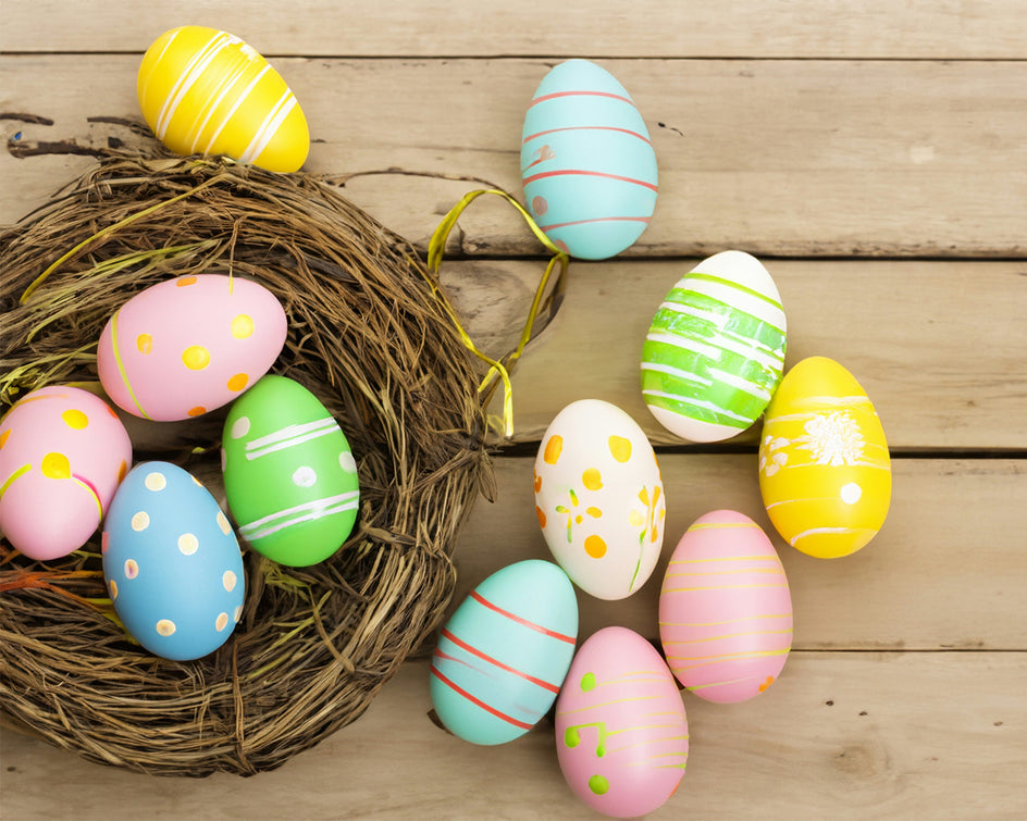 Ein Arrangement aus bunt bemalten Ostereiern in Pastellfarben mit verschiedenen Mustern wie Punkten und Streifen. Die Eier liegen teils in einem dekorativen Nest und teils auf einer Holzoberfläche verteilt.