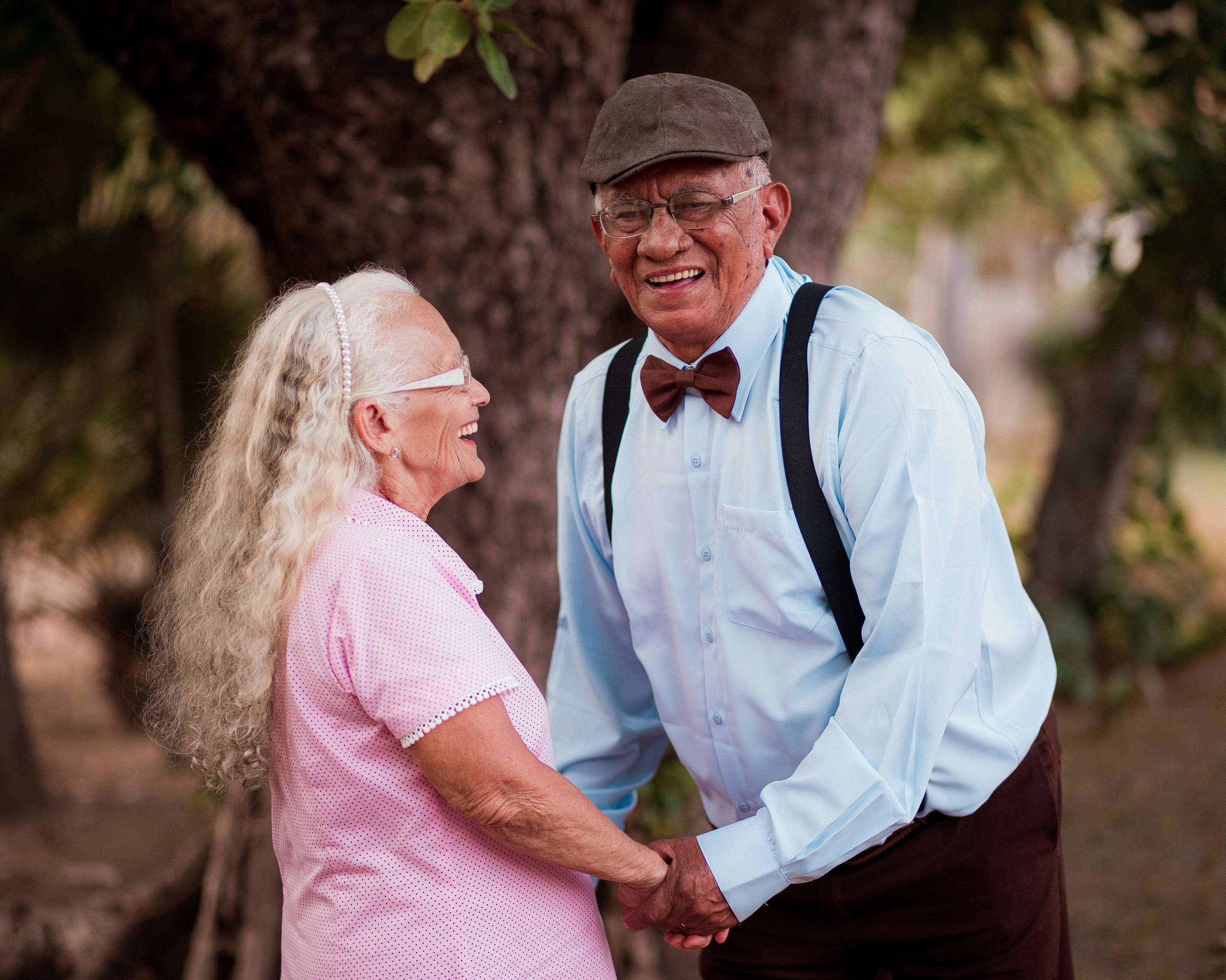 Ein älteres Paar steht im Freien unter einem Baum und hält sich liebevoll an den Händen. Die Frau trägt ein rosa Hemd mit kurzen Ärmeln, während der Mann ein hellblaues Hemd mit Hosenträgern und einer Fliege trägt. Die Szene vermittelt eine warme und harmonische Atmosphäre, umgeben von natürlicher Umgebung.