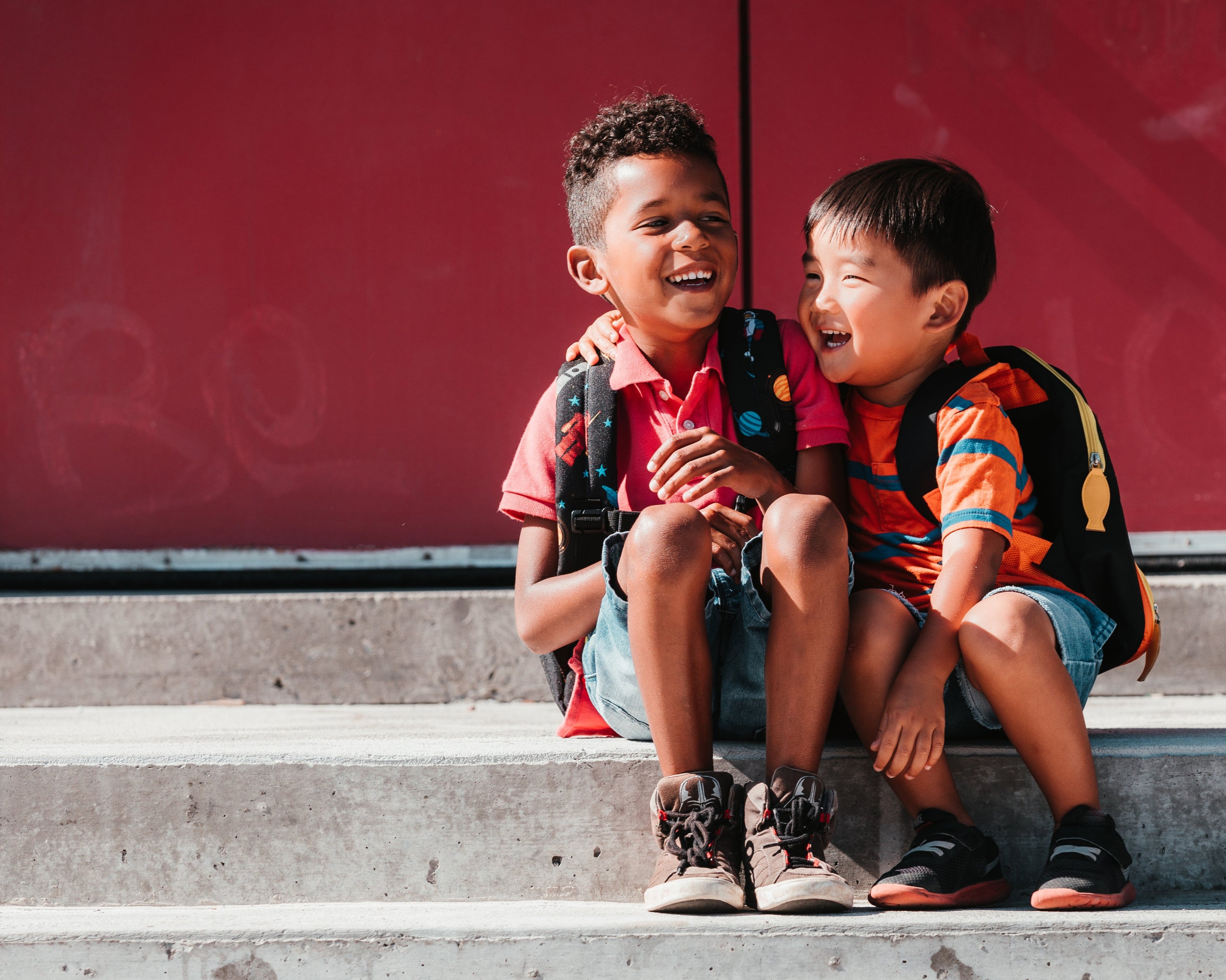 Zwei Kinder sitzen auf einer Treppe vor einer roten Wand. Beide tragen Rucksäcke, bunte T-Shirts und Shorts. Die Szene zeigt eine freundliche Geste, bei der eines der Kinder die Hand auf die Schulter des anderen legt. Die Atmosphäre wirkt warm und sonnig.