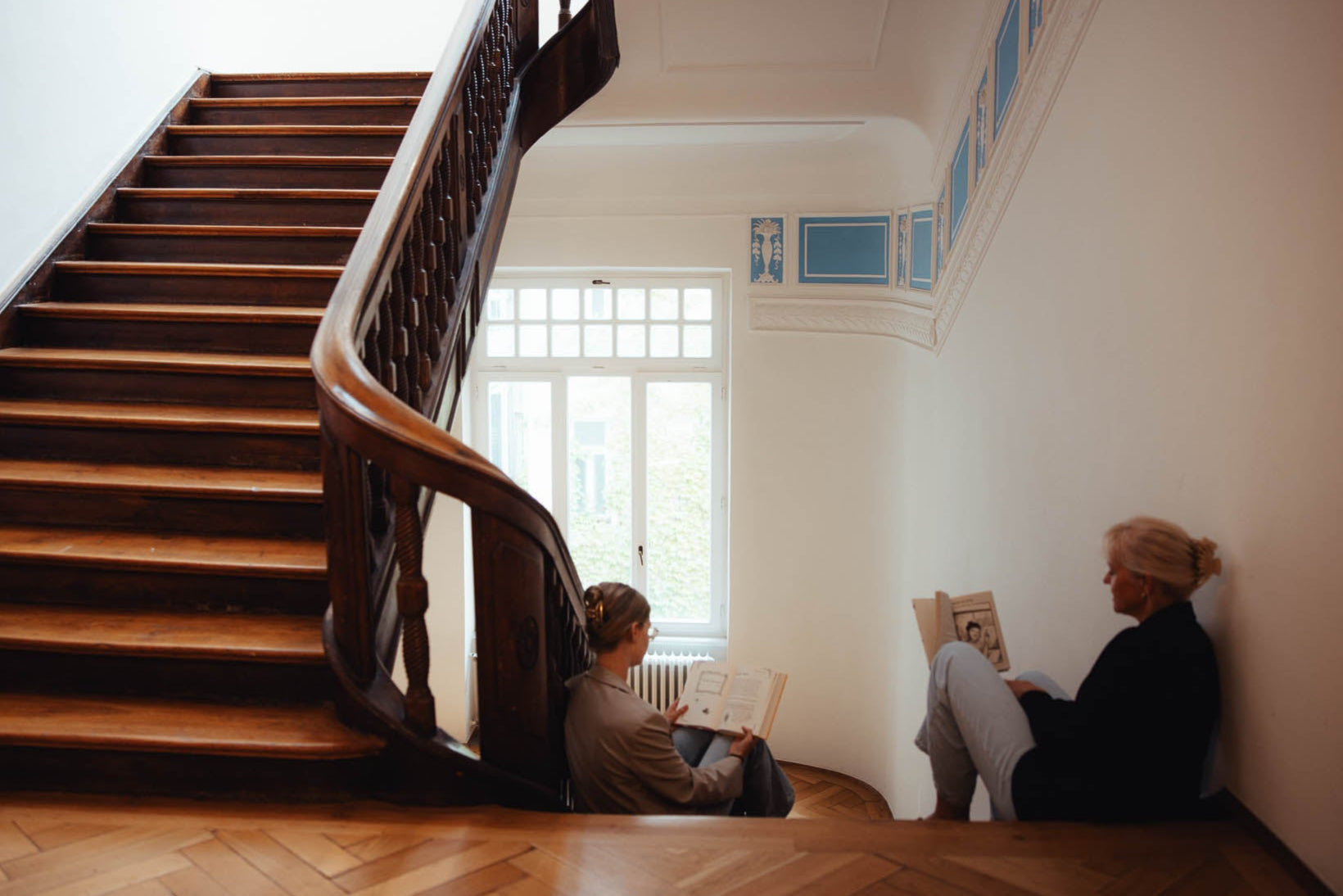 Ein historisches Treppenhaus mit einer dunklen Holztreppe und verzierten blauen Wandornamenten. Zwei Personen sitzen auf dem Boden im Treppenabsatz, vertieft in das Lesen von Büchern. Tageslicht fällt durch ein großes Fenster im Hintergrund, das eine ruhige und gemütliche Atmosphäre schafft.