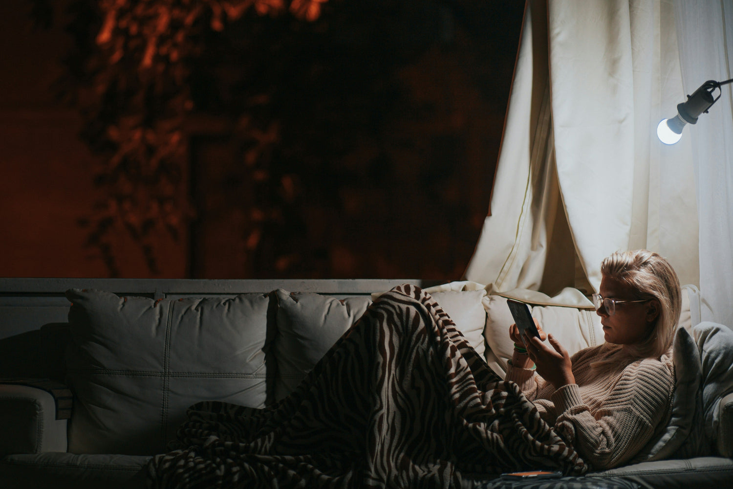 Eine Person sitzt in der Nacht auf einem Sofa unter einer Decke mit Zebramuster und liest auf einem Tablet. Im Hintergrund sind Bäume und eine Hausfassade im warmen Licht der Straßenbeleuchtung zu sehen. Eine Lampe an der Wand beleuchtet die Szene dezent.