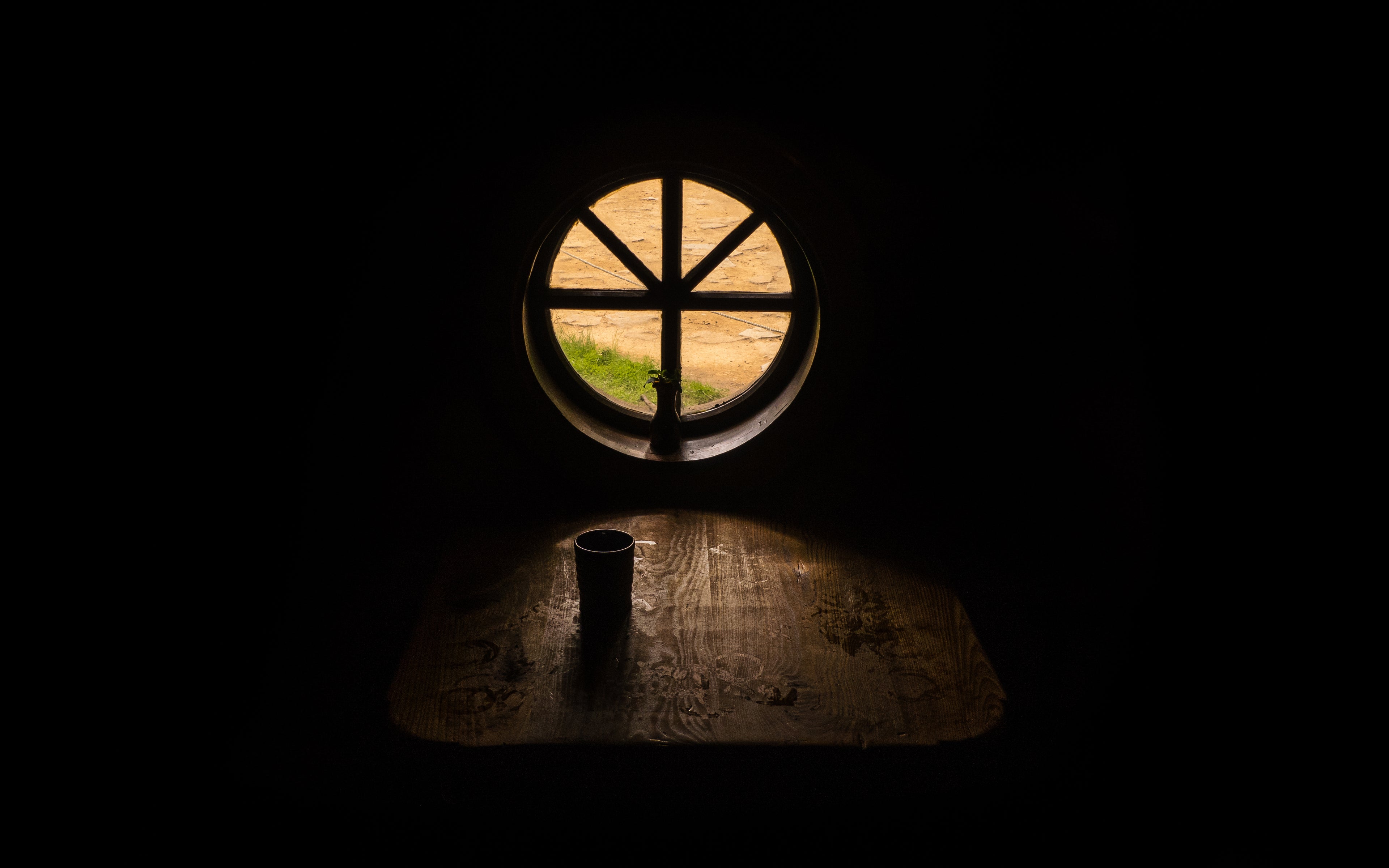 Ein rundes Fenster mit einem Holzrahmen, durch das Tageslicht auf einen rustikalen Holztisch fällt. Auf dem Tisch steht ein dunkler Becher, und im Hintergrund ist ein sandiger Weg mit etwas Grün zu sehen. Die Umgebung des Raums ist dunkel, wodurch das Licht und die Details des Fensters und Tisches hervorgehoben werden.