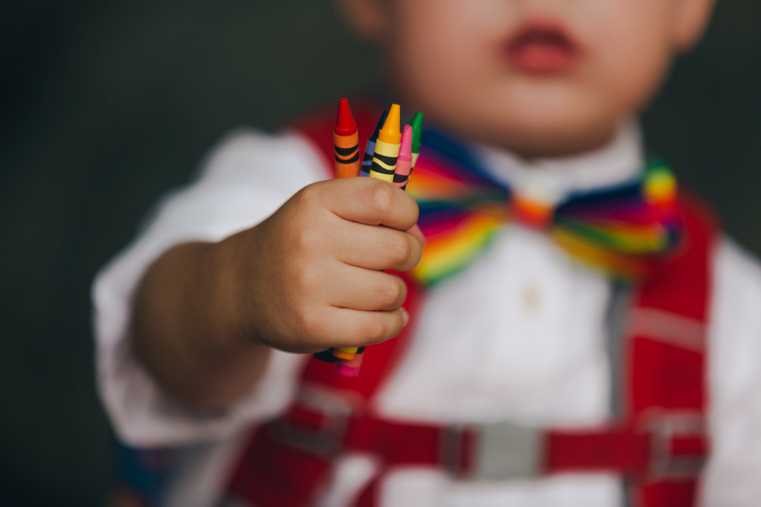 Ein Kleinkind hält eine Handvoll bunter Wachsmalstifte in den Farben Rot, Gelb, Grün und Pink. Im Hintergrund ist ein weißes Hemd mit einer regenbogenfarbenen Fliege und roten Hosenträgern zu sehen. Der Fokus liegt auf der ausgestreckten Hand mit den Stiften, während das Gesicht des Kindes unscharf ist.