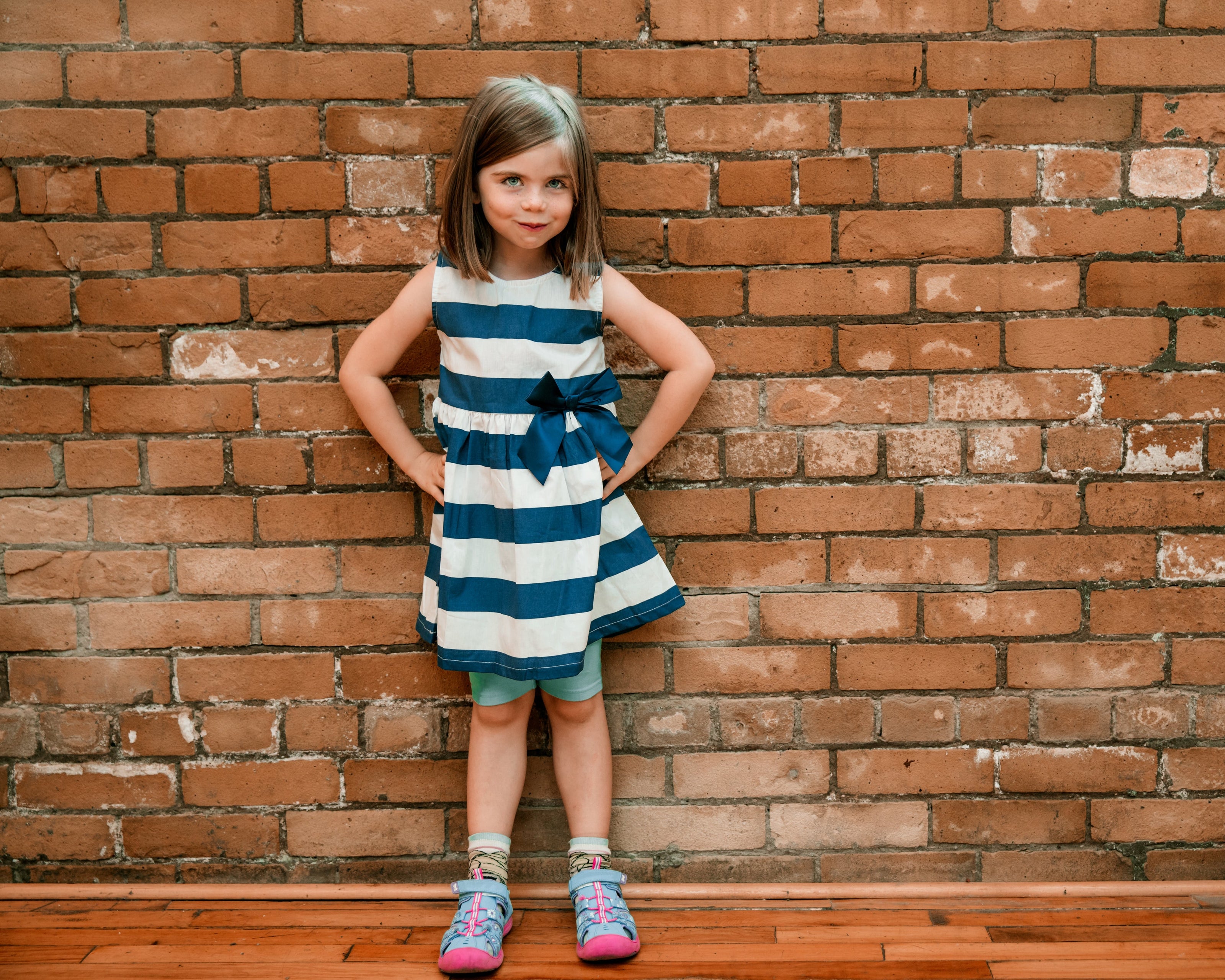 Ein Kind steht vor einer roten Backsteinwand und trägt ein weiß-blau gestreiftes Kleid mit einer Schleife in der Taille. Dazu kombiniert es bunte Schuhe und grüne Leggings. Die Hände sind in die Hüften gestützt, und der Hintergrund zeigt eine strukturierte Ziegelsteinoberfläche.