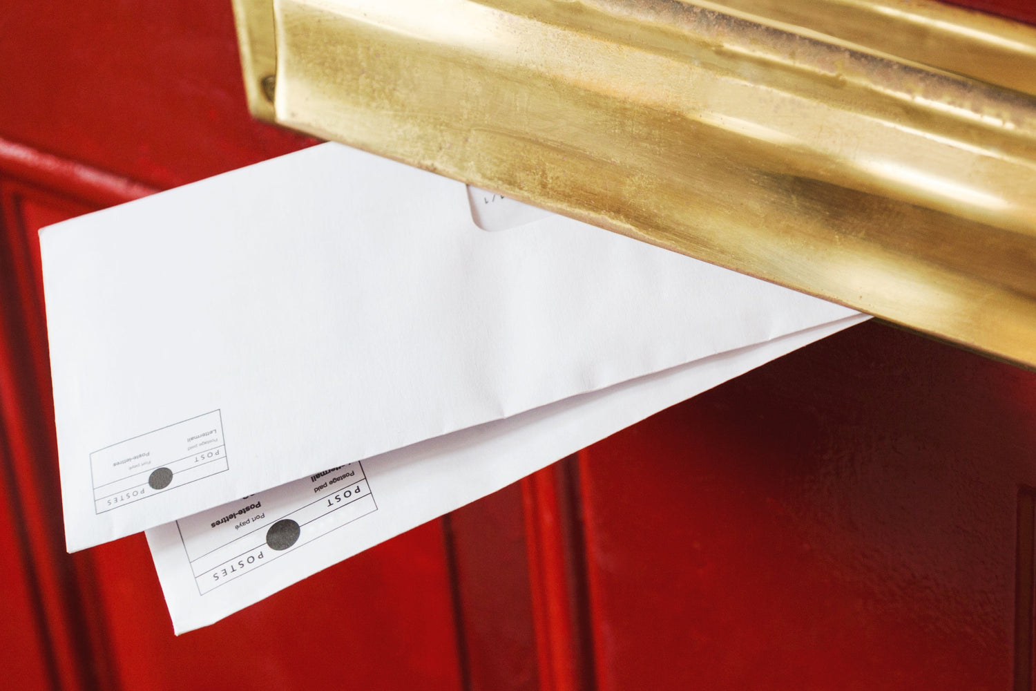 Ein roter Türbereich mit einem goldfarbenen Briefschlitz, aus dem zwei weiße Umschläge herausragen. Die Umschläge haben Postmarkierungen und scheinen teilweise eingeworfen zu sein.