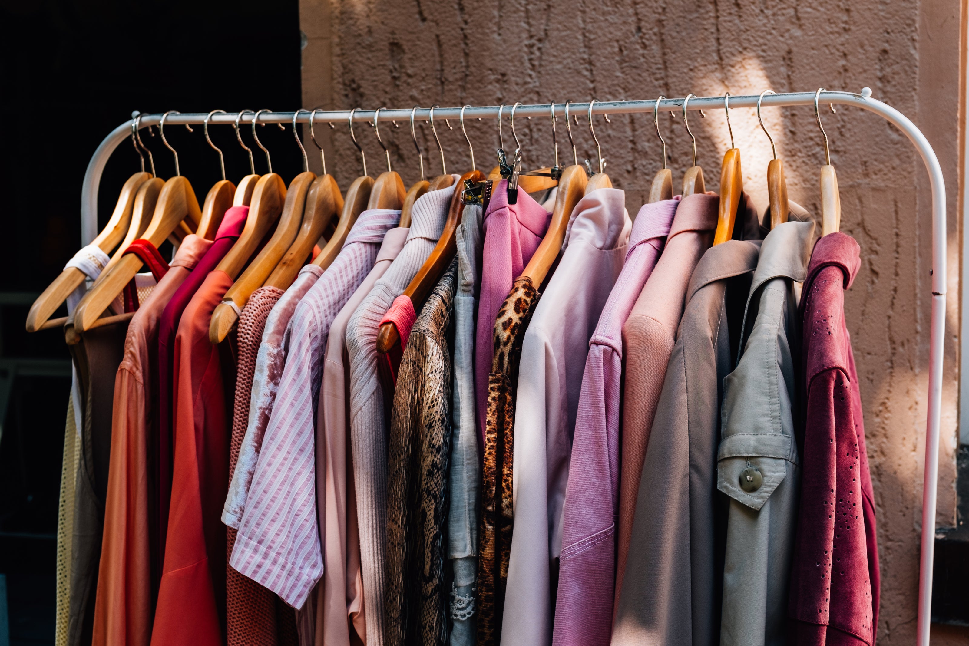 Ein Kleiderständer mit hölzernen Kleiderbügeln, an denen verschiedene Kleidungsstücke in warmen Farbtönen wie Rosa, Rot und Beige hängen. Die Kleidung umfasst Hemden, Blusen und Jacken, die ordentlich nebeneinander angeordnet sind. Der Hintergrund zeigt eine grobe, beige Wand.