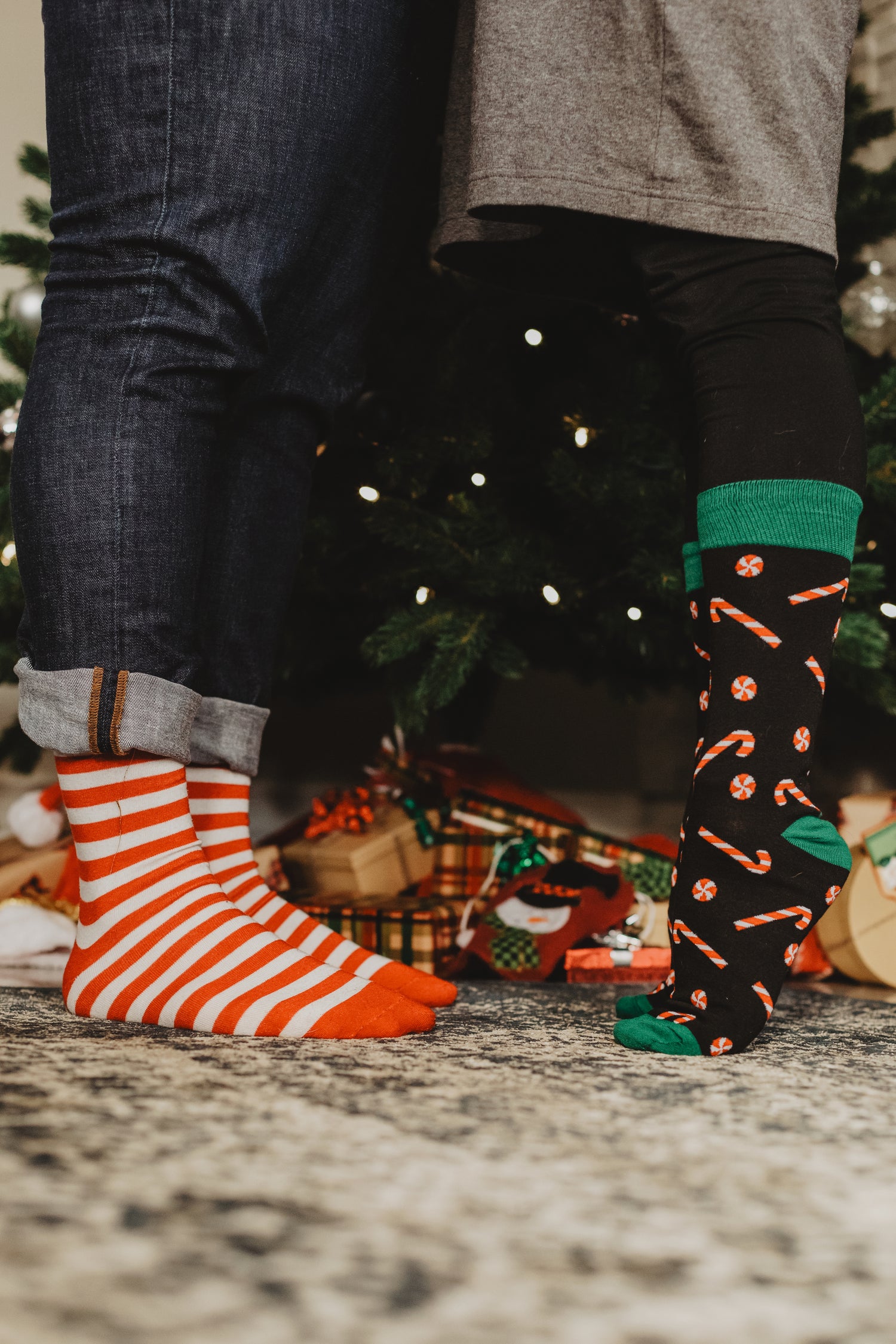 Zwei Personen stehen nebeneinander vor einem geschmückten Weihnachtsbaum. Die linke Person trägt rot-weiß gestreifte Socken und Jeans, während die rechte Person schwarze Socken mit Zuckerstangen-Muster und grünem Bündchen trägt. Im Hintergrund sind Geschenke unter dem Baum sichtbar.