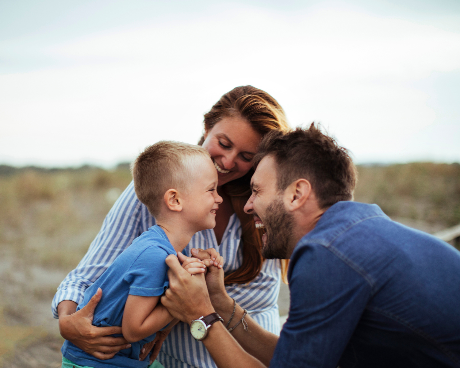 Ein Junge im blauen T-Shirt wird von zwei Erwachsenen liebevoll umarmt. Die Szene spielt im Freien mit unscharfem Hintergrund aus Gras und Himmel. Der Mann rechts trägt ein blaues Jeanshemd und eine Armbanduhr, die Frau links trägt ein gestreiftes Oberteil.