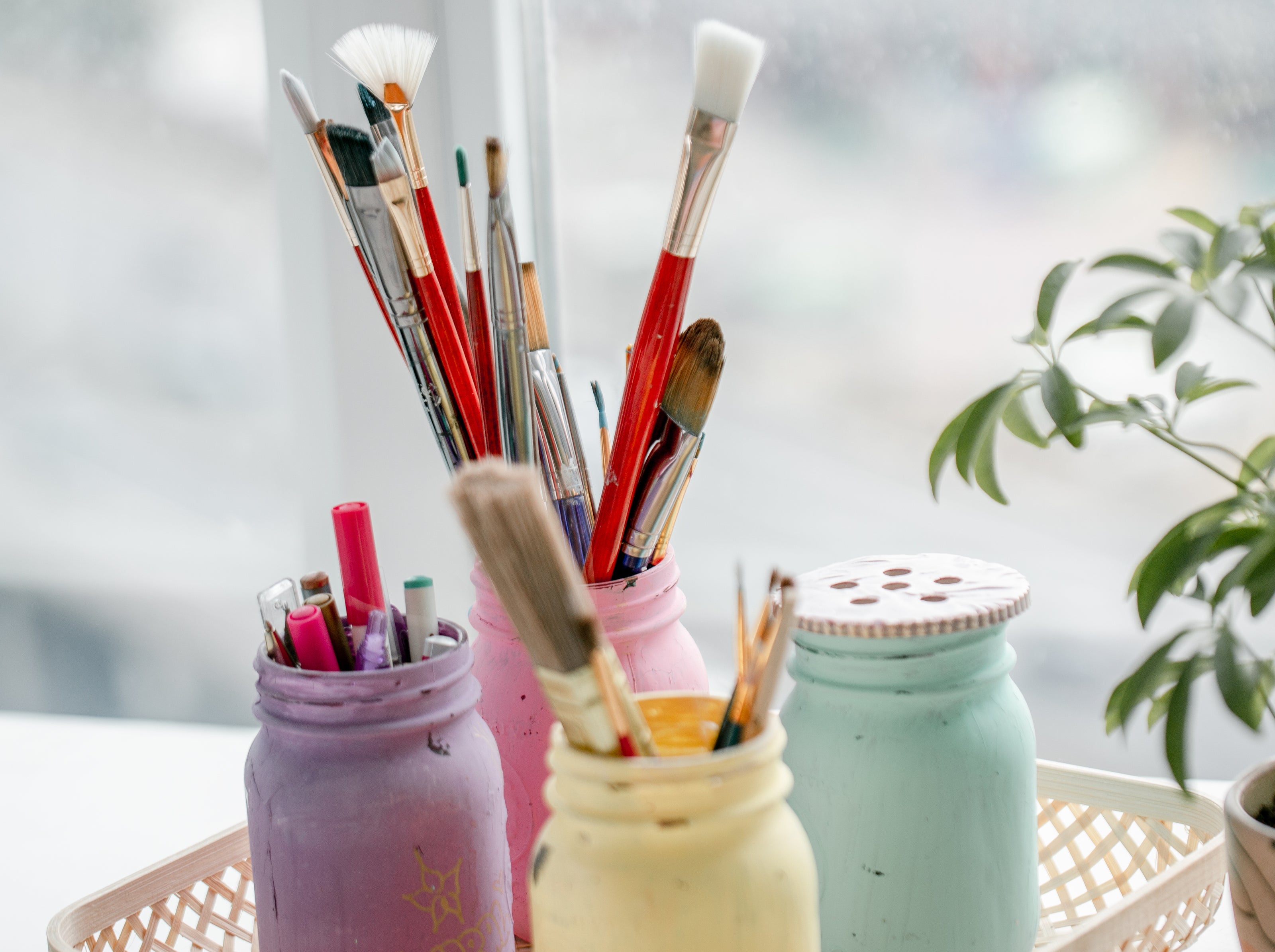 Verschiedene Pinsel und Stifte stehen in bemalten Gläsern in Pastellfarben (lila, rosa, mintgrün und hellgelb) auf einem hellen Tisch vor einem Fenster. Im Hintergrund ist unscharf eine Pflanze zu sehen.