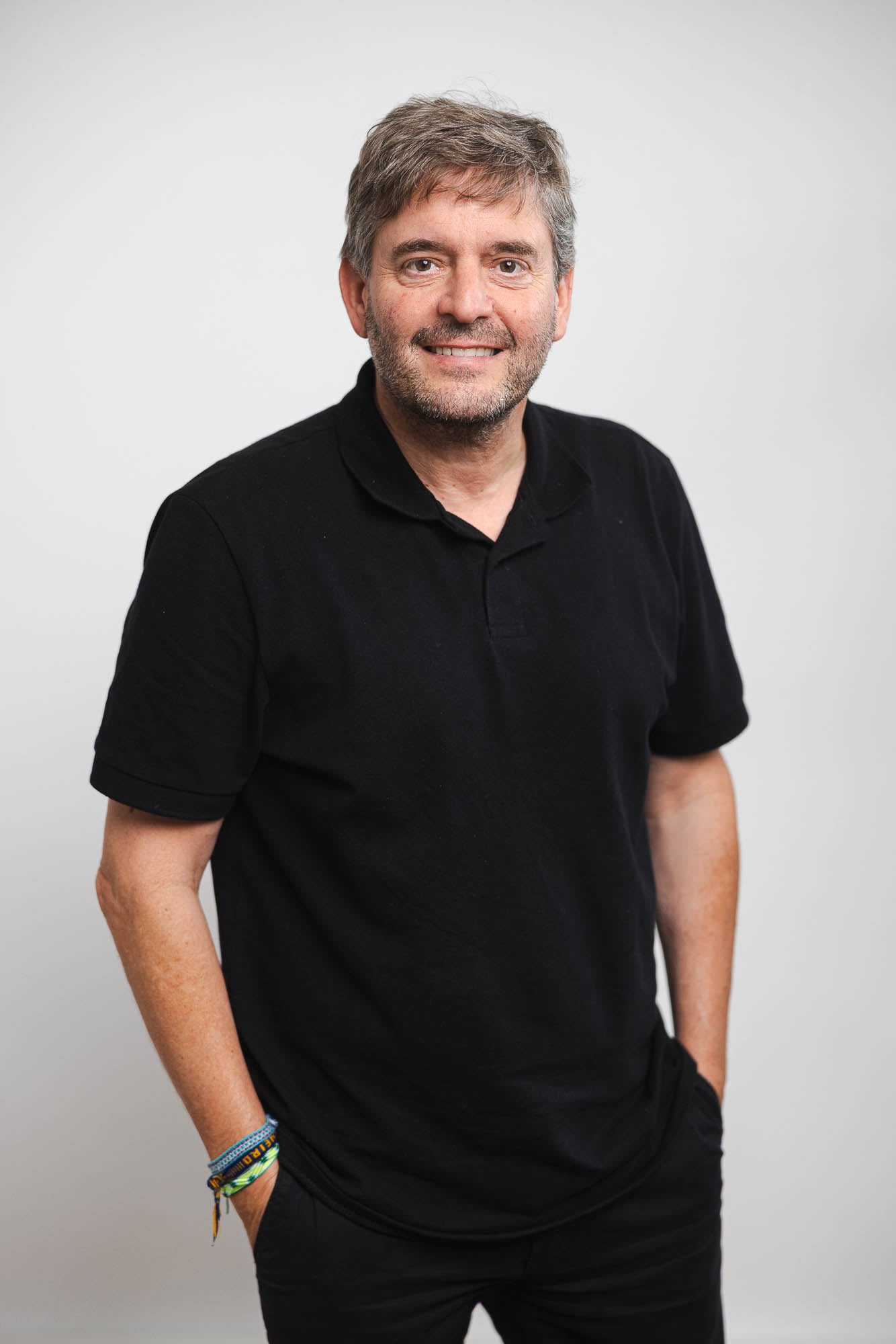 Ralph Benkel steht vor einem neutralen, weißen Hintergrund. Er trägt ein schwarzes Poloshirt, eine schwarze Hose und mehrere bunte Armbänder am linken Handgelenk.