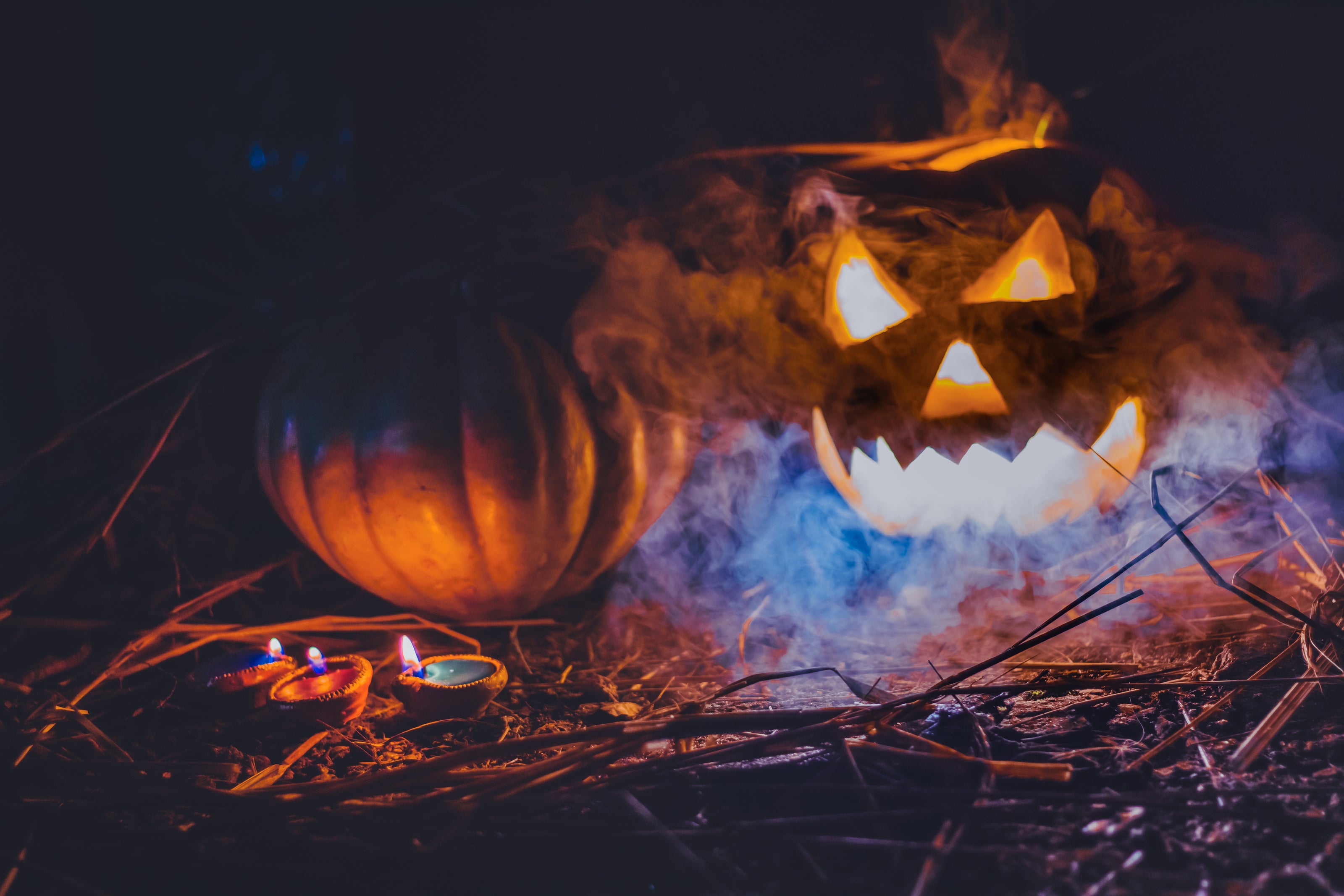 Ein beleuchteter Halloween-Kürbis mit gruseligem Gesicht, aus dem Rauch aufsteigt. Daneben steht ein weiterer unbeleuchteter Kürbis. Im Vordergrund befinden sich kleine, brennende Teelichter, umgeben von trockenen Zweigen und einem dunklen, mystischen Hintergrund.