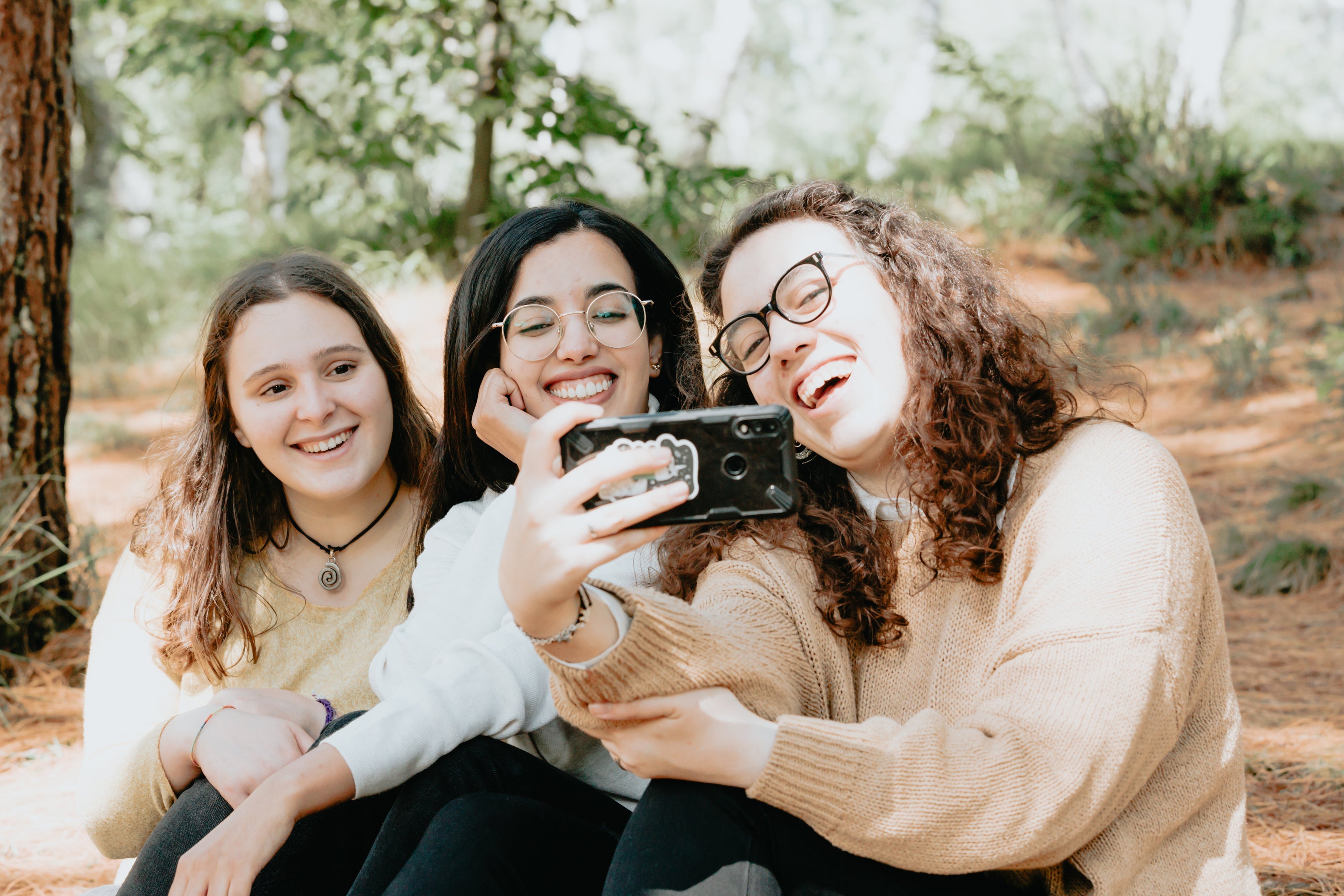 Eine Gruppe von drei Personen sitzt im Freien auf dem Boden eines Waldgebiets und macht ein Selfie mit einem Smartphone. Im Hintergrund sind Bäume und natürliche Vegetation zu sehen.