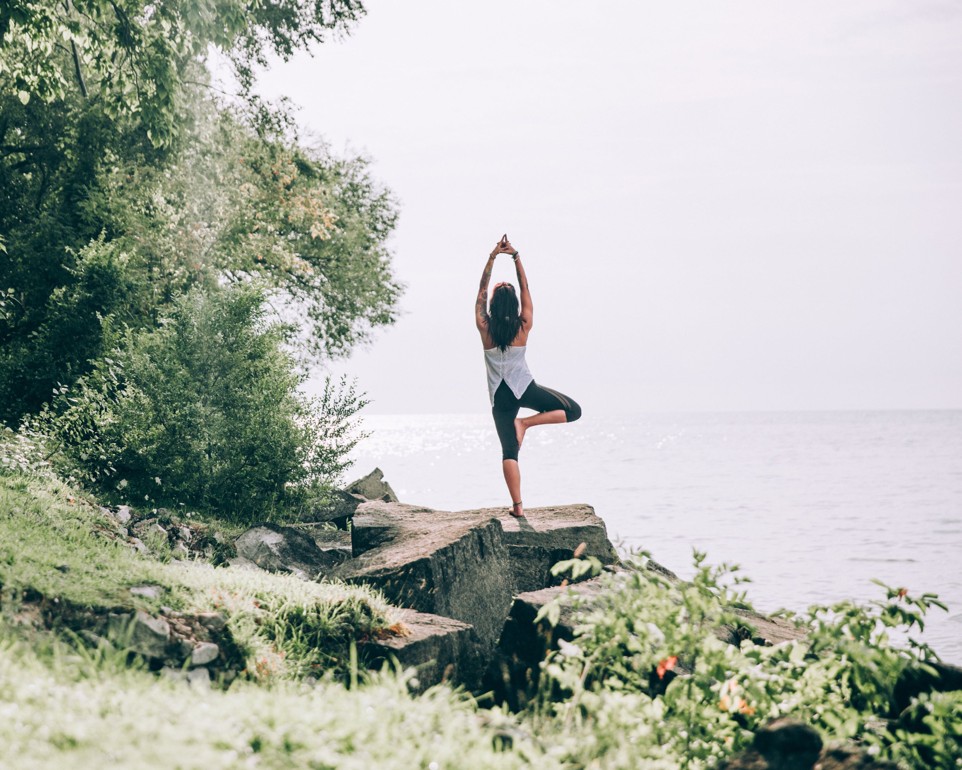 Eine Frau steht in der Yoga-Baumhaltung auf einem Felsen am Ufer eines Sees. Sie trägt sportliche Kleidung und hebt die Arme über den Kopf. Im Hintergrund sind Bäume und das ruhige Wasser des Sees zu sehen, die eine friedliche, natürliche Atmosphäre schaffen.