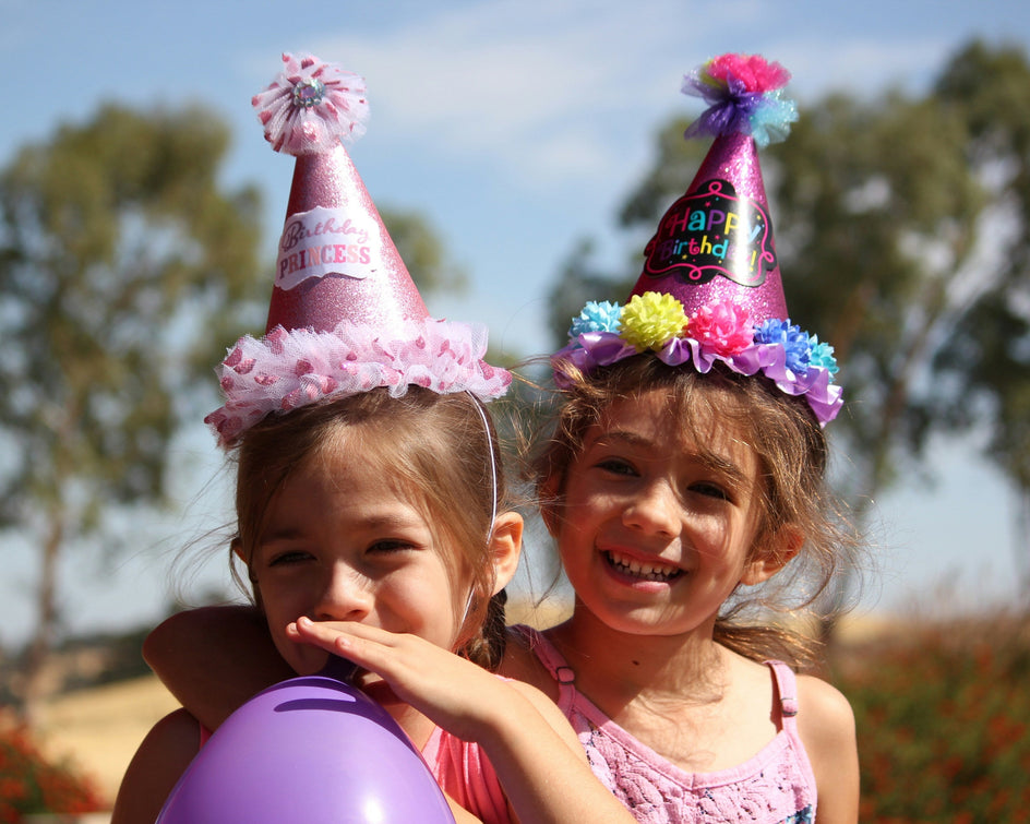 Zwei Kinder tragen glitzernde Partyhüte mit Geburtstagsmotiven. Eines hält einen lilafarbenen Ballon. Im Hintergrund sind Bäume und ein sonniger Himmel zu sehen, die auf eine Feier im Freien hindeuten.