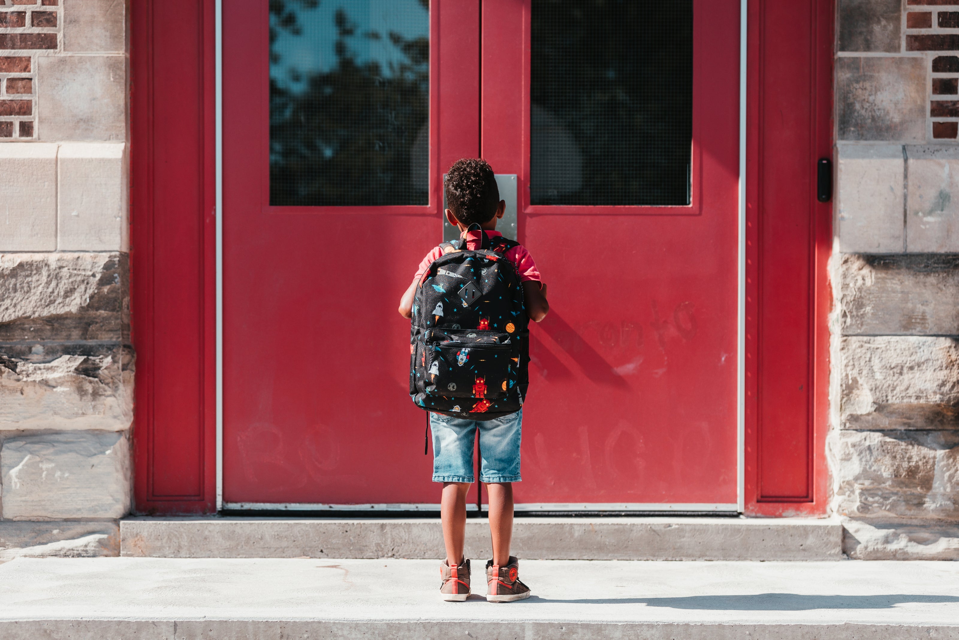 Ein Kind mit lockigem Haar steht mit dem Rücken zur Kamera vor einer roten Tür aus Metall und Glas. Es trägt einen schwarzen Rucksack mit buntem Muster, ein rotes T-Shirt, Jeans-Shorts und braune Schuhe. Die Tür gehört zu einem Gebäude aus Stein.