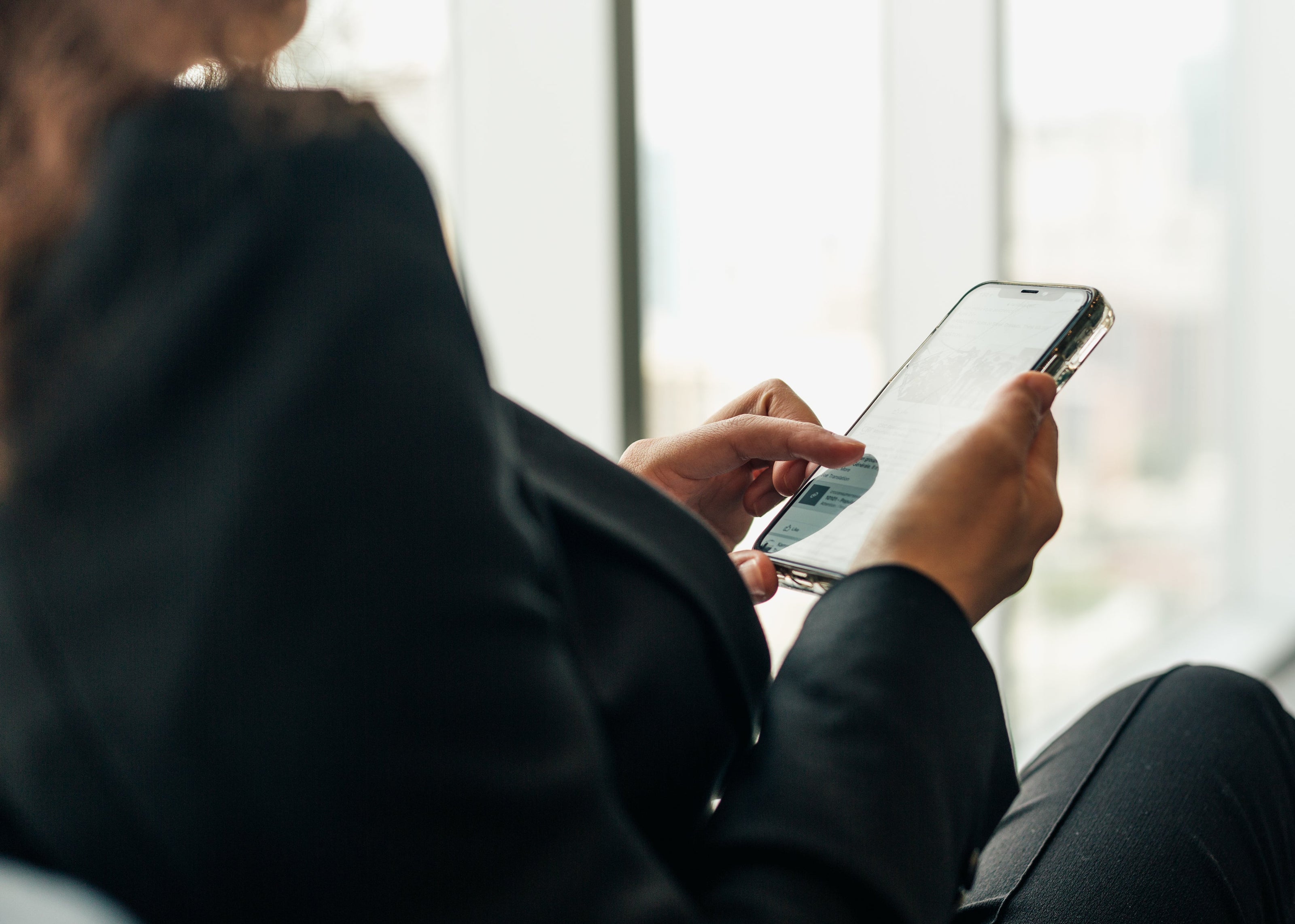 Eine Person in dunkler Kleidung hält ein Smartphone in der Hand und betrachtet den Bildschirm. Im Hintergrund sind unscharf Fenster mit Blick auf eine Stadtlandschaft erkennbar.