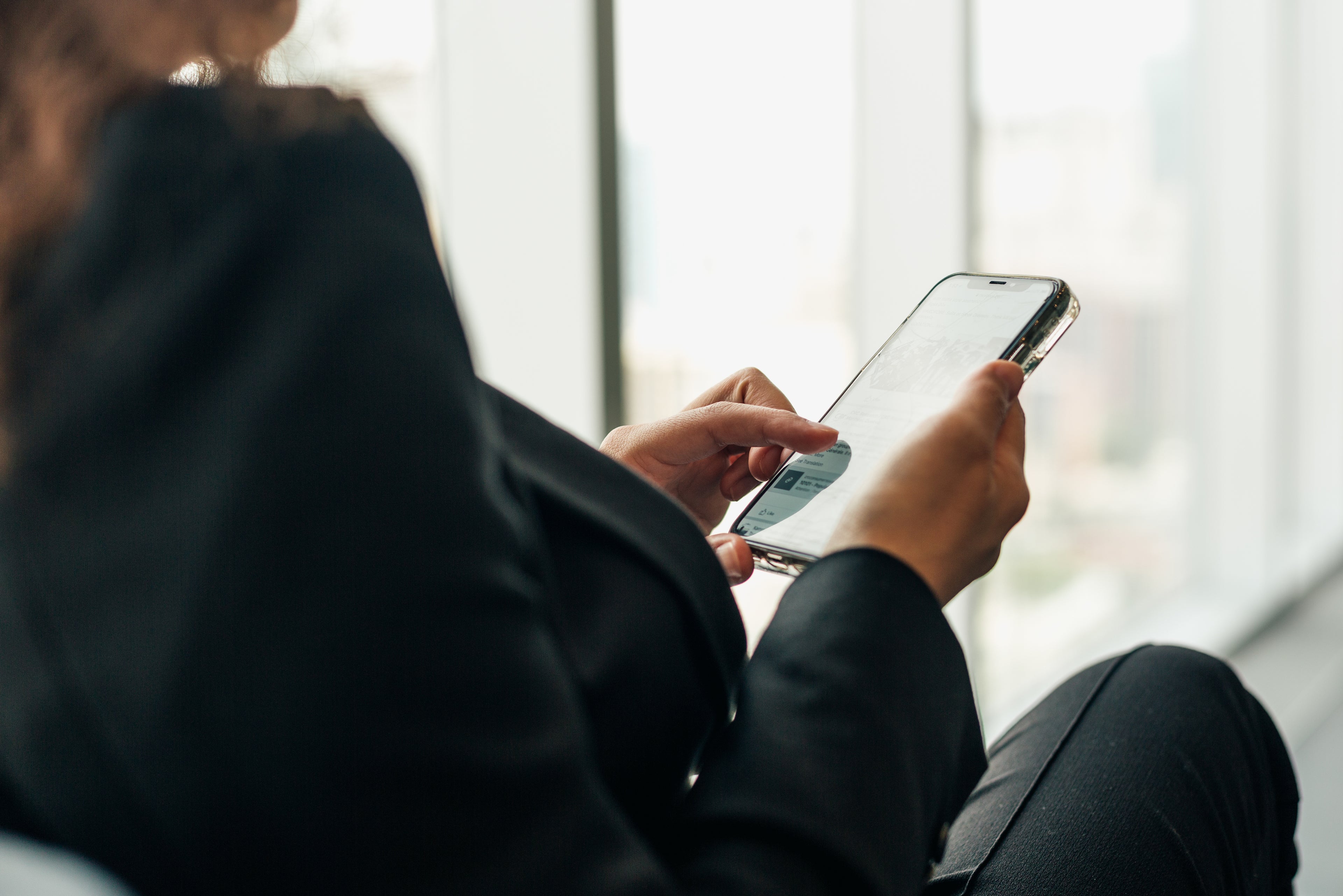 Gezeigt wird eine Person in dunkler Kleidung, die ein Smartphone in beiden Händen hält und mit dem Finger auf dem Bildschirm tippt. Das Gerät wird in Nahaufnahme gezeigt, während der Hintergrund mit großen Fenstern und hellem Tageslicht unscharf bleibt.