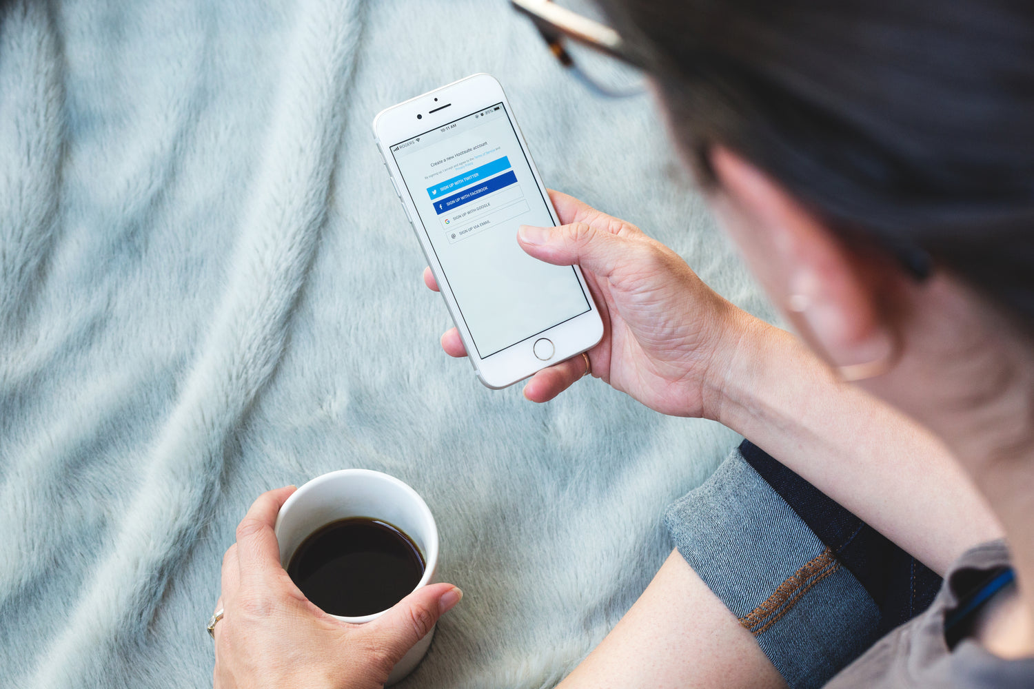Das Foto zeigt eine Person von oben, die auf einer hellen, weichen Decke sitzt. Sie hält in einer Hand eine weiße Tasse mit Kaffee und in der anderen ein Smartphone. Auf dem Display ist eine Anmeldeseite mit mehreren Schaltflächen für verschiedene Registrierungsoptionen zu sehen.