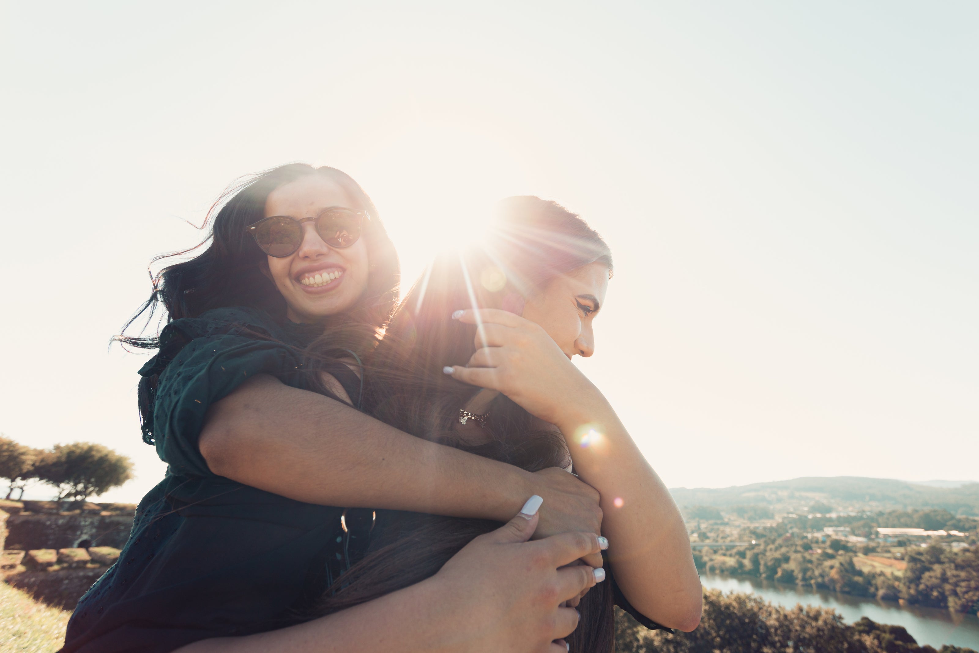 Zwei Frauen umarmen sich im Freien bei Sonnenuntergang. Der Hintergrund zeigt eine malerische Landschaft mit Hügeln, Wasser und Bäumen, während die Sonne strahlend zwischen ihnen hindurchscheint.