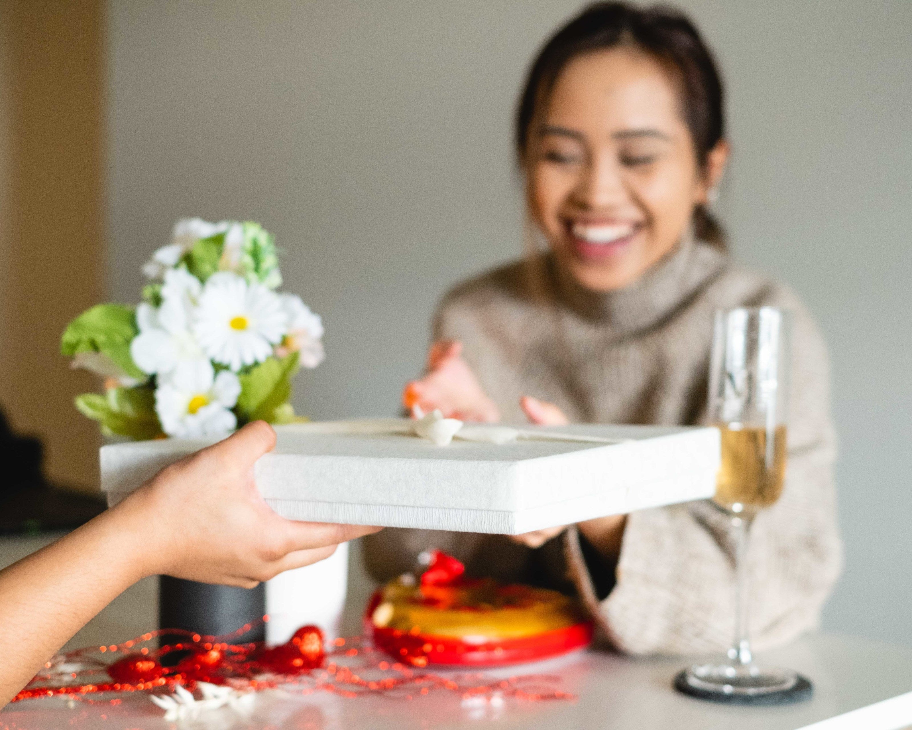 A close-up image shows a hand offering a gift box with a small decorative flower on top. In the background, there is a vase with white daisies, a champagne glass, and festive red decorations on a table.