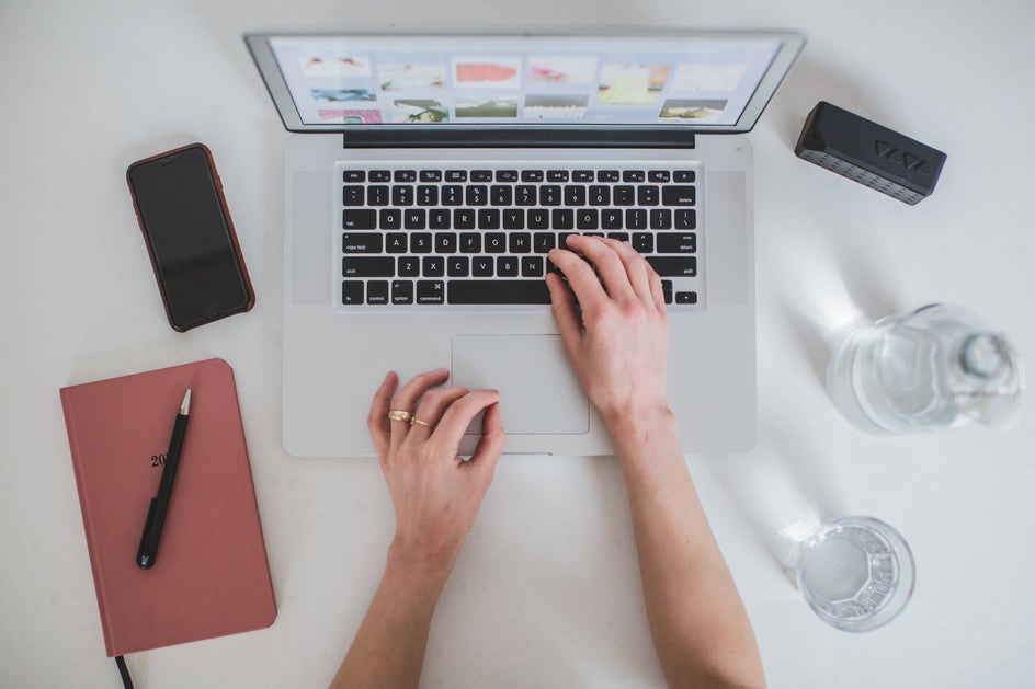 Sicht von oben auf einen Arbeitsplatz. Zwei Frauenhände tippen auf einem Laptop. Links liegt ein Smartphone und Notizbuch, rechts stehen Wasserflasche und Glas.