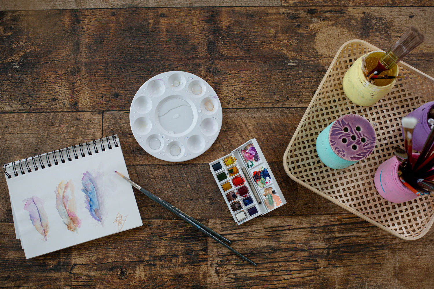 Arbeitsplatz für Aquarellmalerei mit Skizzenbuch, Farbkasten, Pinseln und Zubehör auf einem Holztisch.