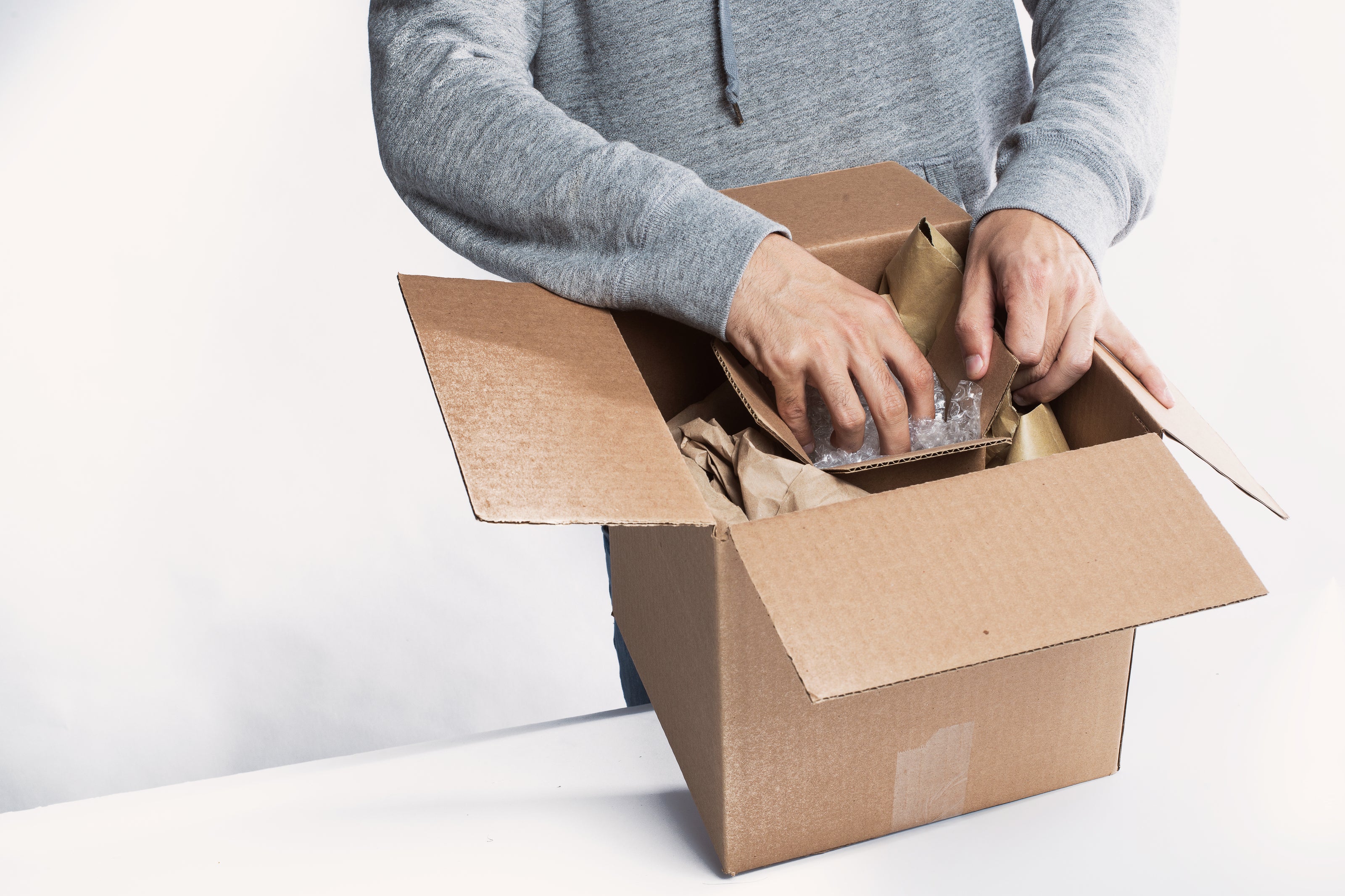 Eine Person in grauem Hoodie packt einen Gegenstand aus einer offenen Pappschachtel aus, umgeben von Verpackungsmaterial. Der Hintergrund ist weiß